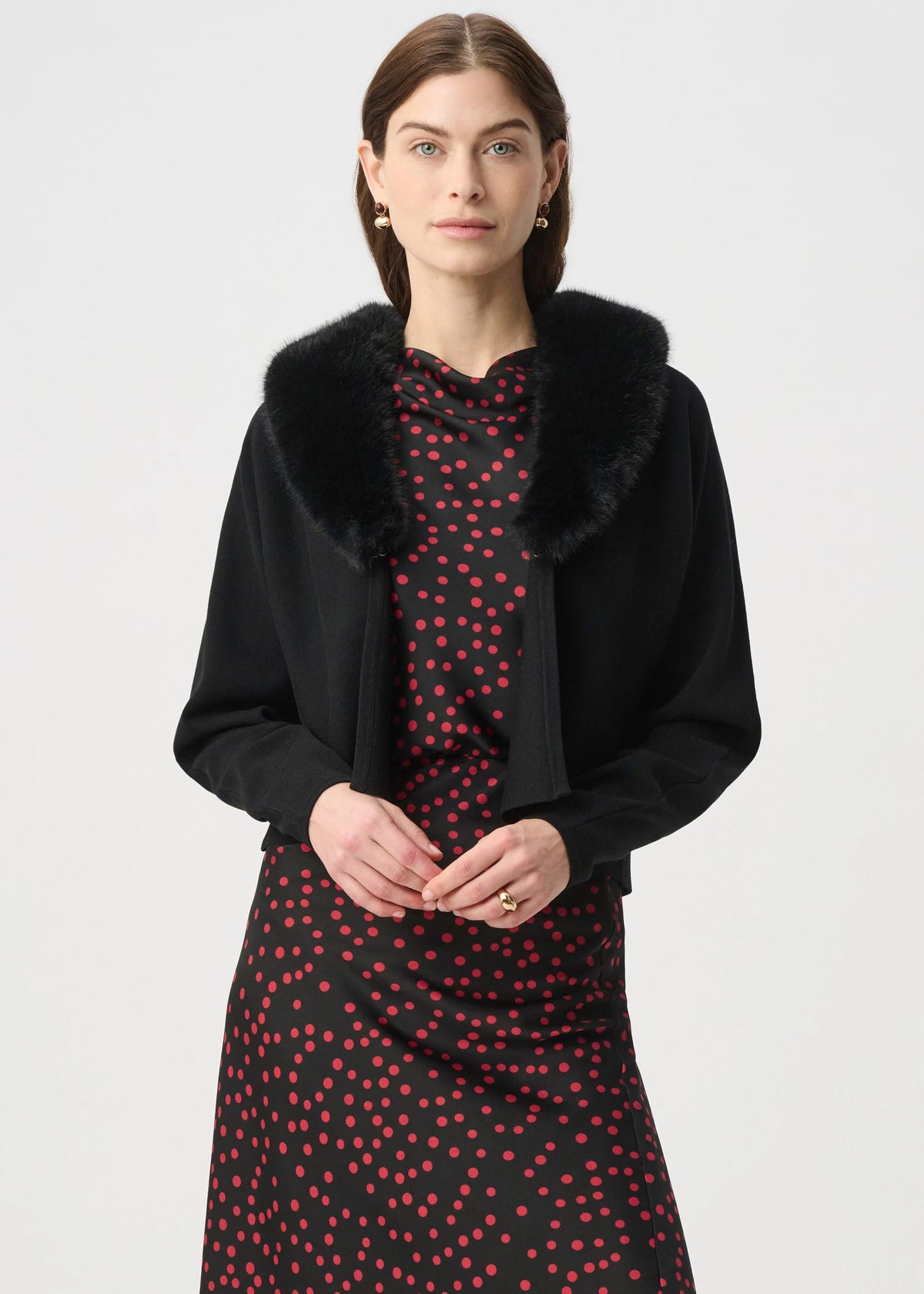 A woman modeling a black long-sleeve jacket with a plush black faux-fur collar, worn over a black dress with a red polka-dot pattern, against a light gray studio background.