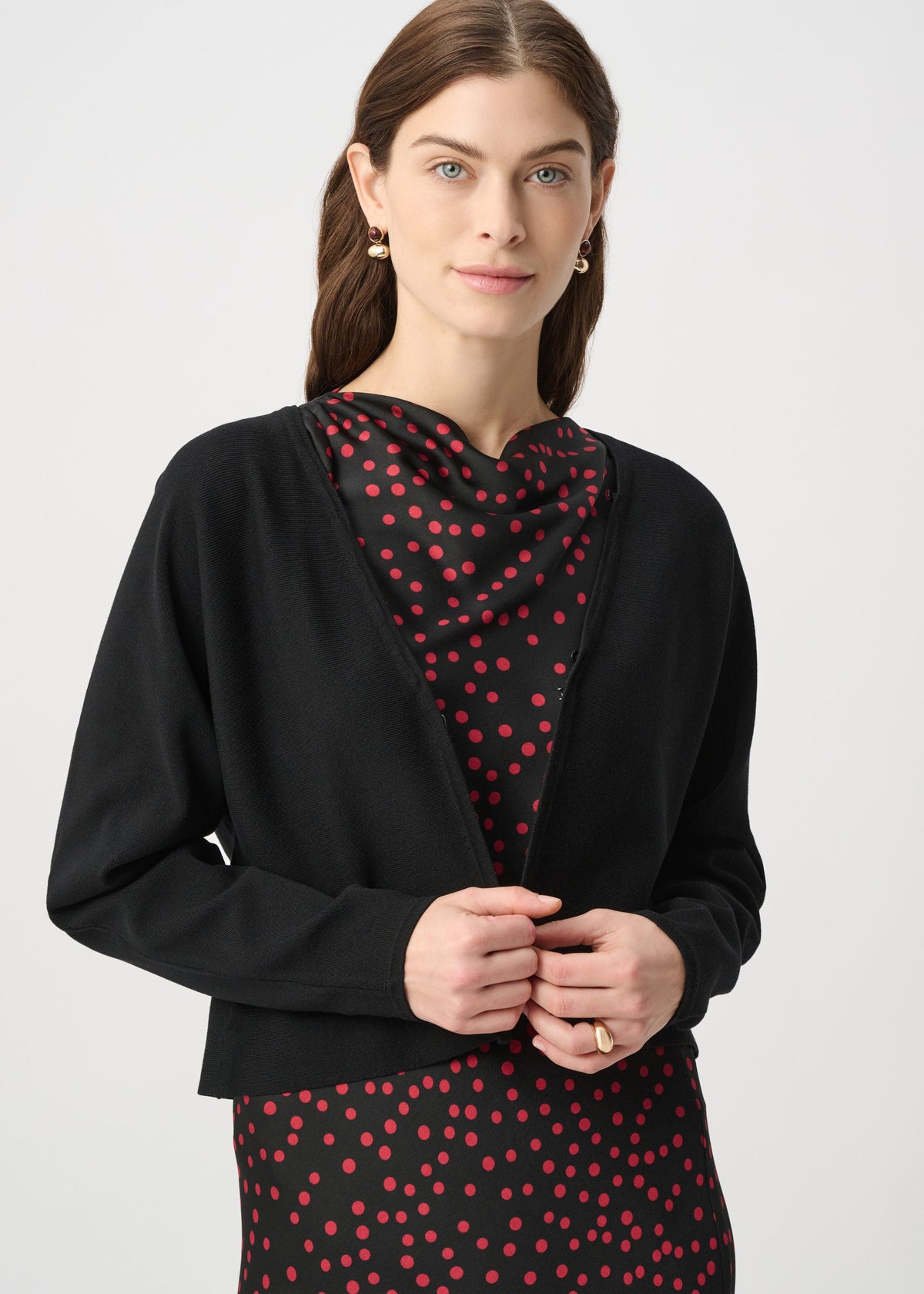 A woman modeling a black cardigan over a black dress with red polka dots, photographed against a light gray studio background.