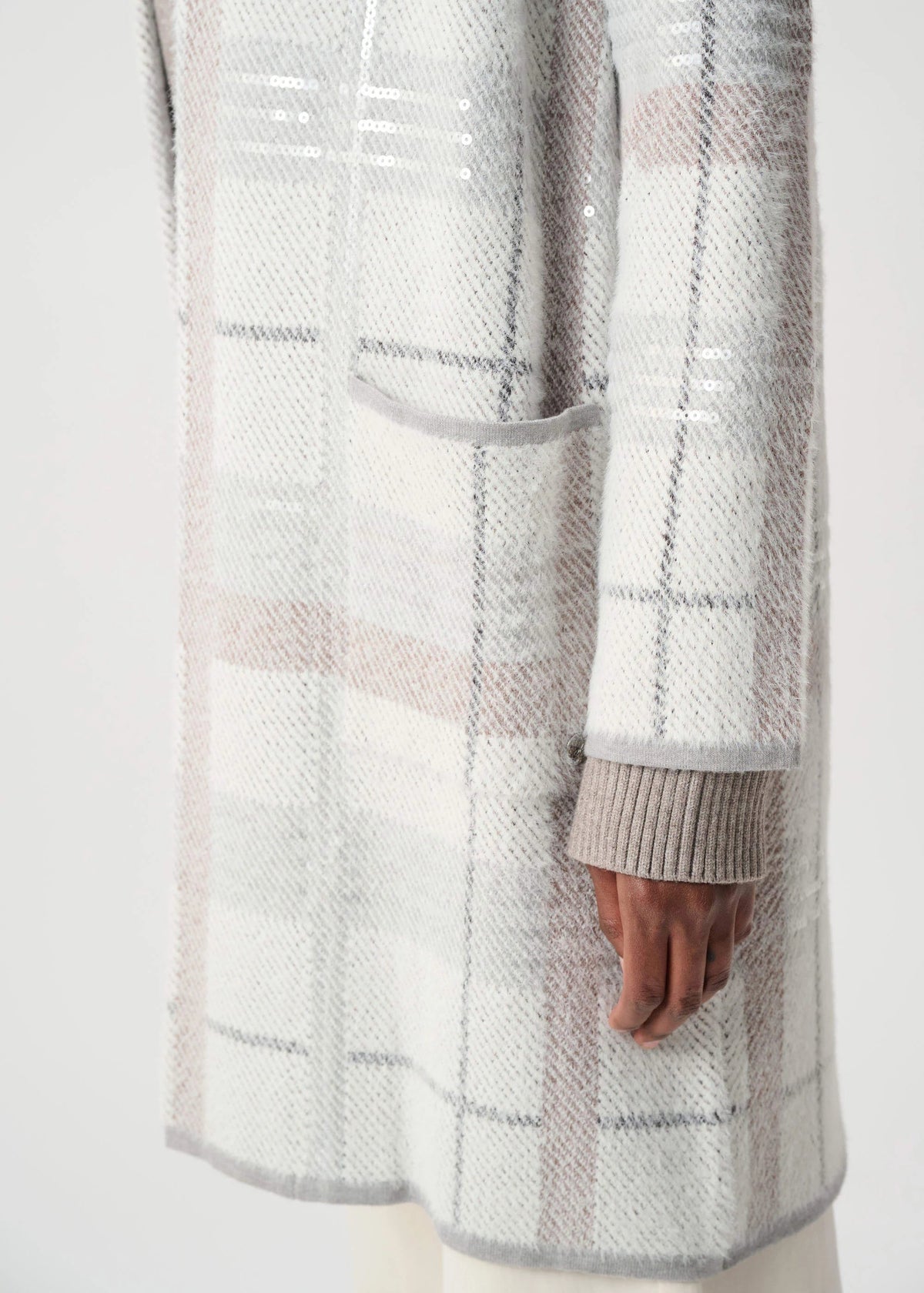 A close-up of a person wearing a textured knit cardigan with a soft plaid pattern in off-white, light gray, and pale pink. The cardigan features a ribbed cuff and a front patch pocket, shown against a plain light background.
