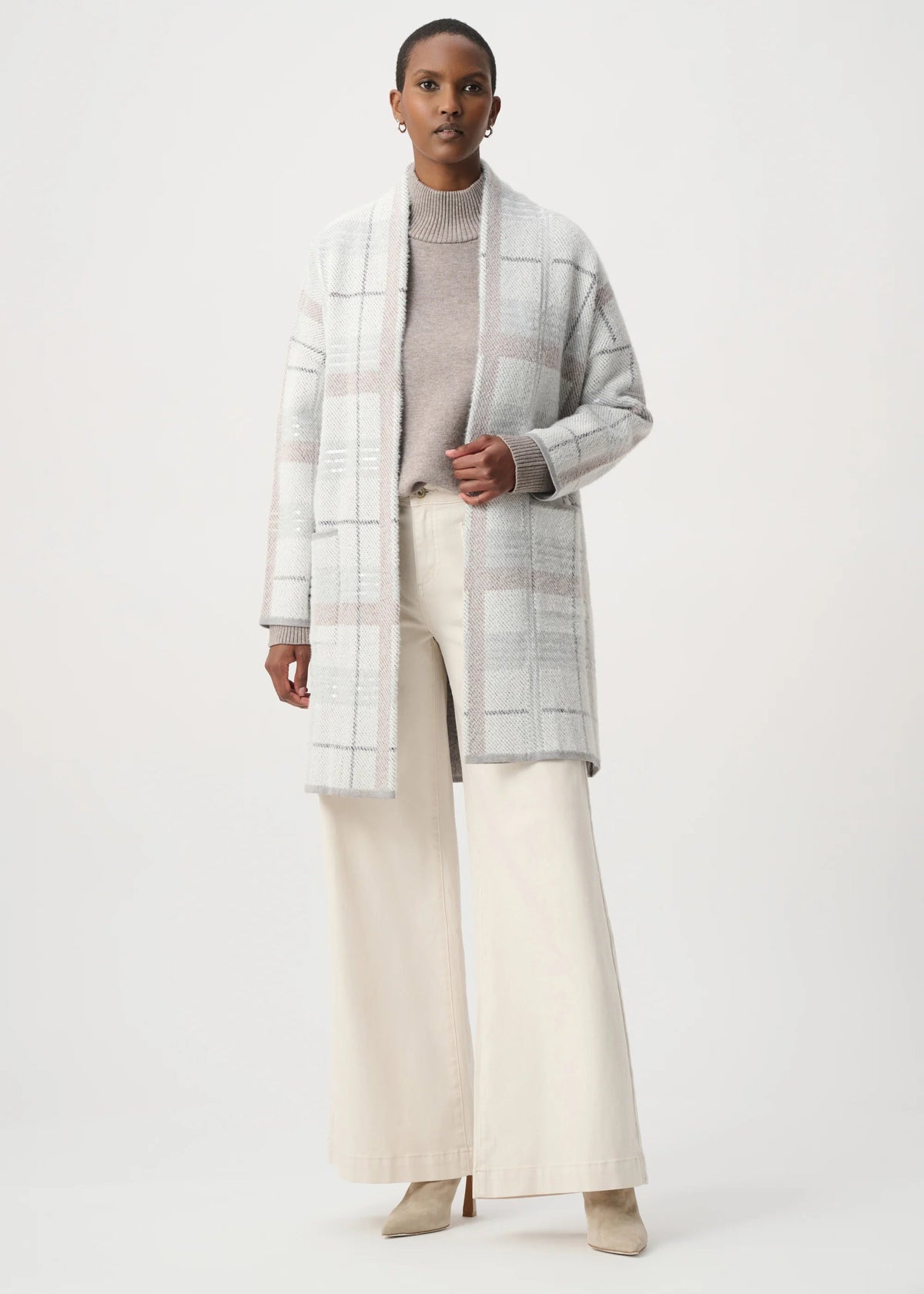 A model stands against a plain light gray studio background wearing a long open-front plaid coat in soft gray, white, and pale pink over a taupe turtleneck, cream wide-leg pants, and beige pointed shoes.