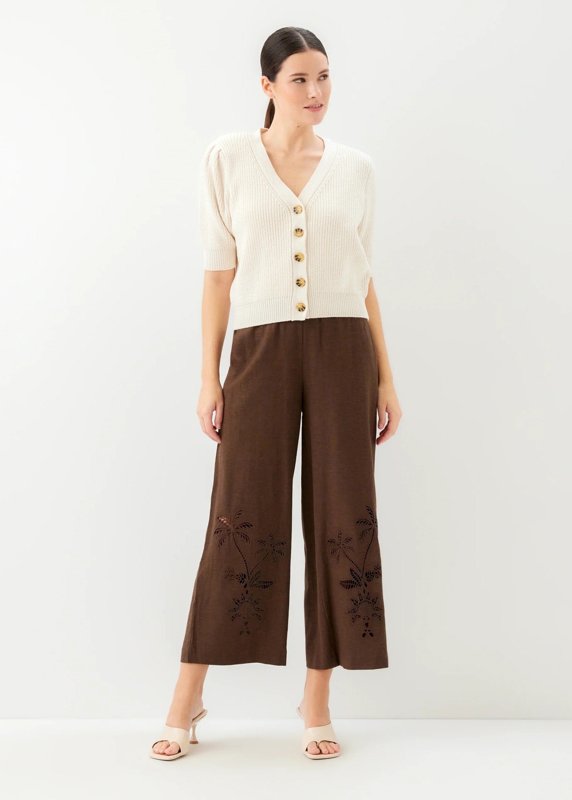 Woman wearing a cream cardigan and brown pants with floral embroidery on a white background
