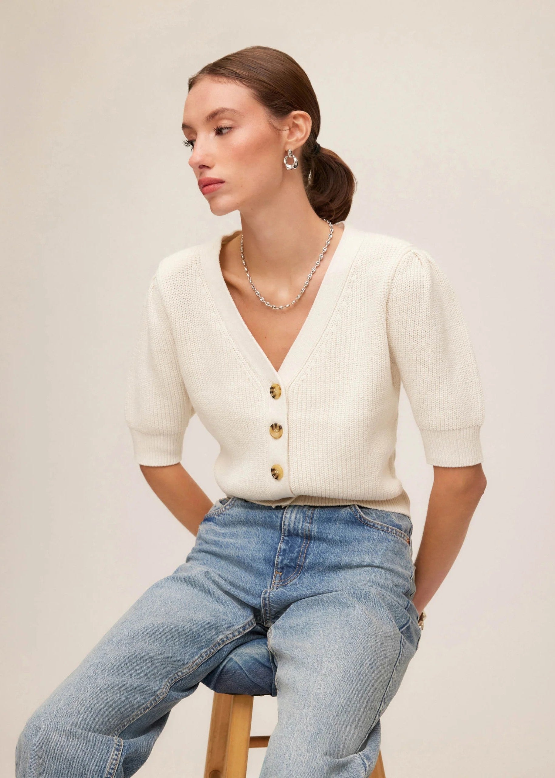 Woman wearing a white cardigan with gold buttons and blue jeans on a plain background