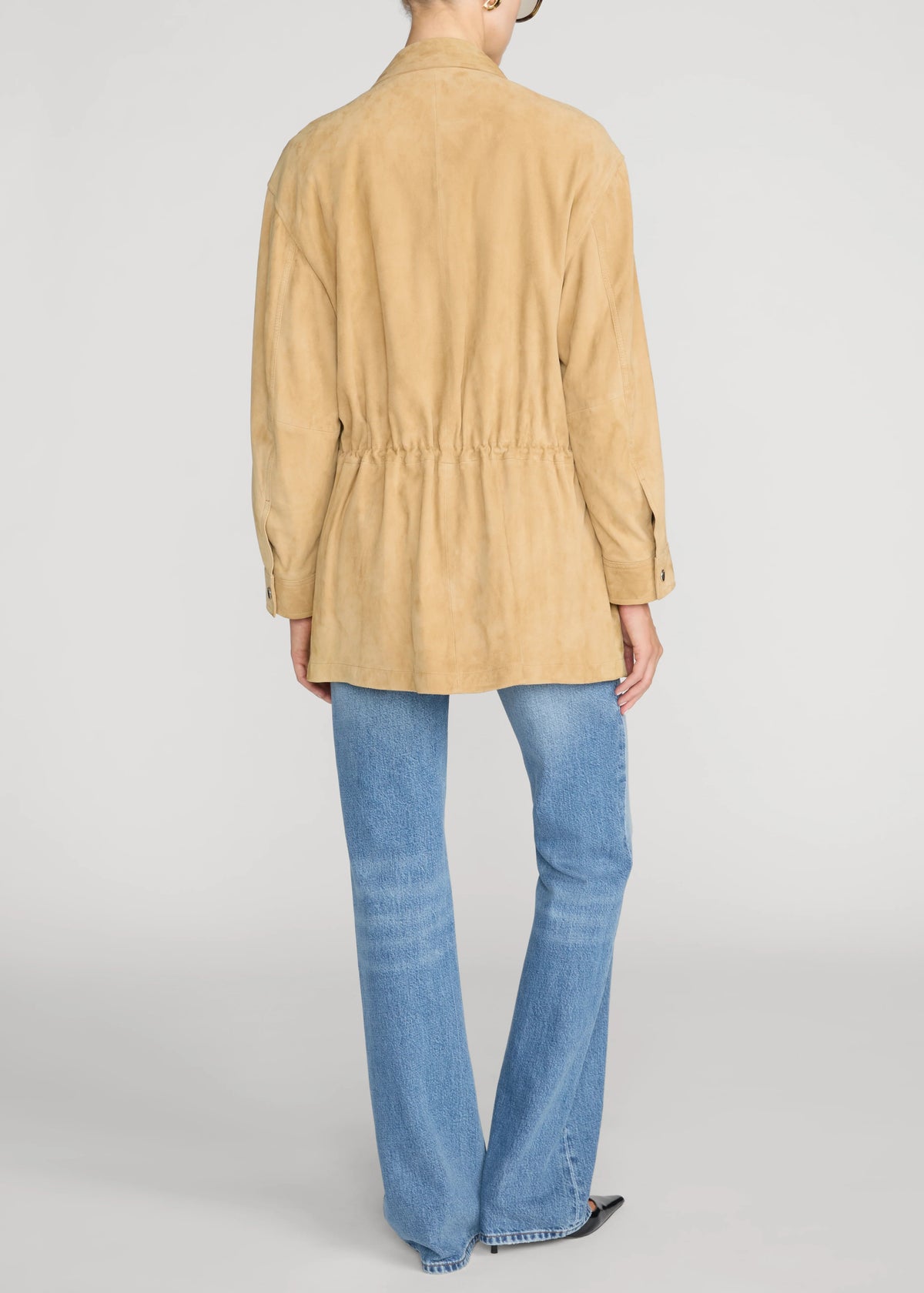 A model shown from the back wearing a tan suede long-sleeve peplum-style jacket over light blue flared jeans, photographed against a neutral light gray studio background.