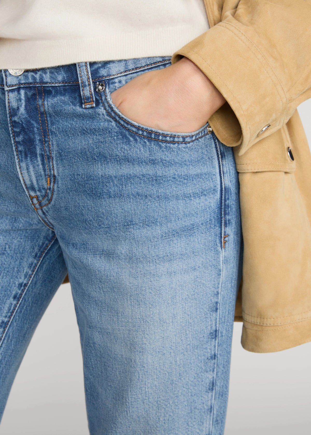 Close-up of a person wearing light blue denim jeans with a hand in the pocket, styled with a cream top and a tan jacket against a light gray background.