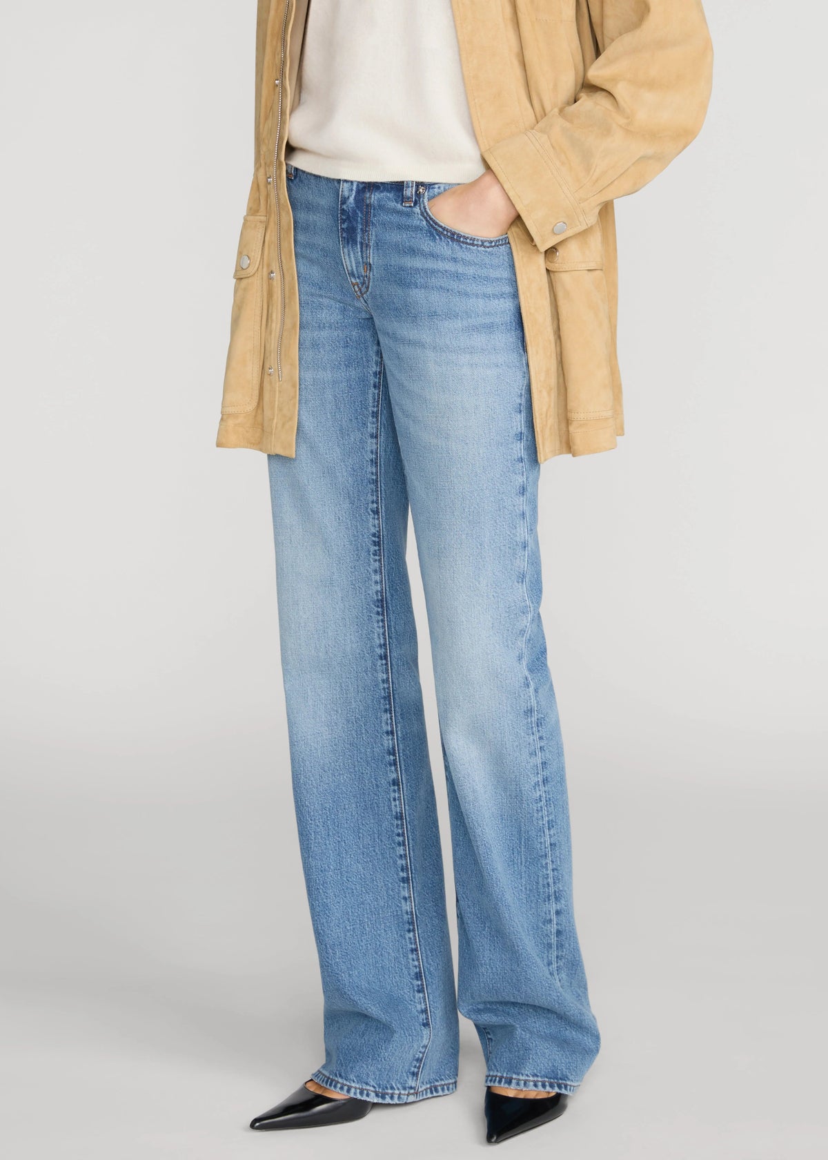 A person wearing light blue wide-leg jeans with a cream top, a tan overshirt, and black pointed shoes, photographed against a light gray studio background.