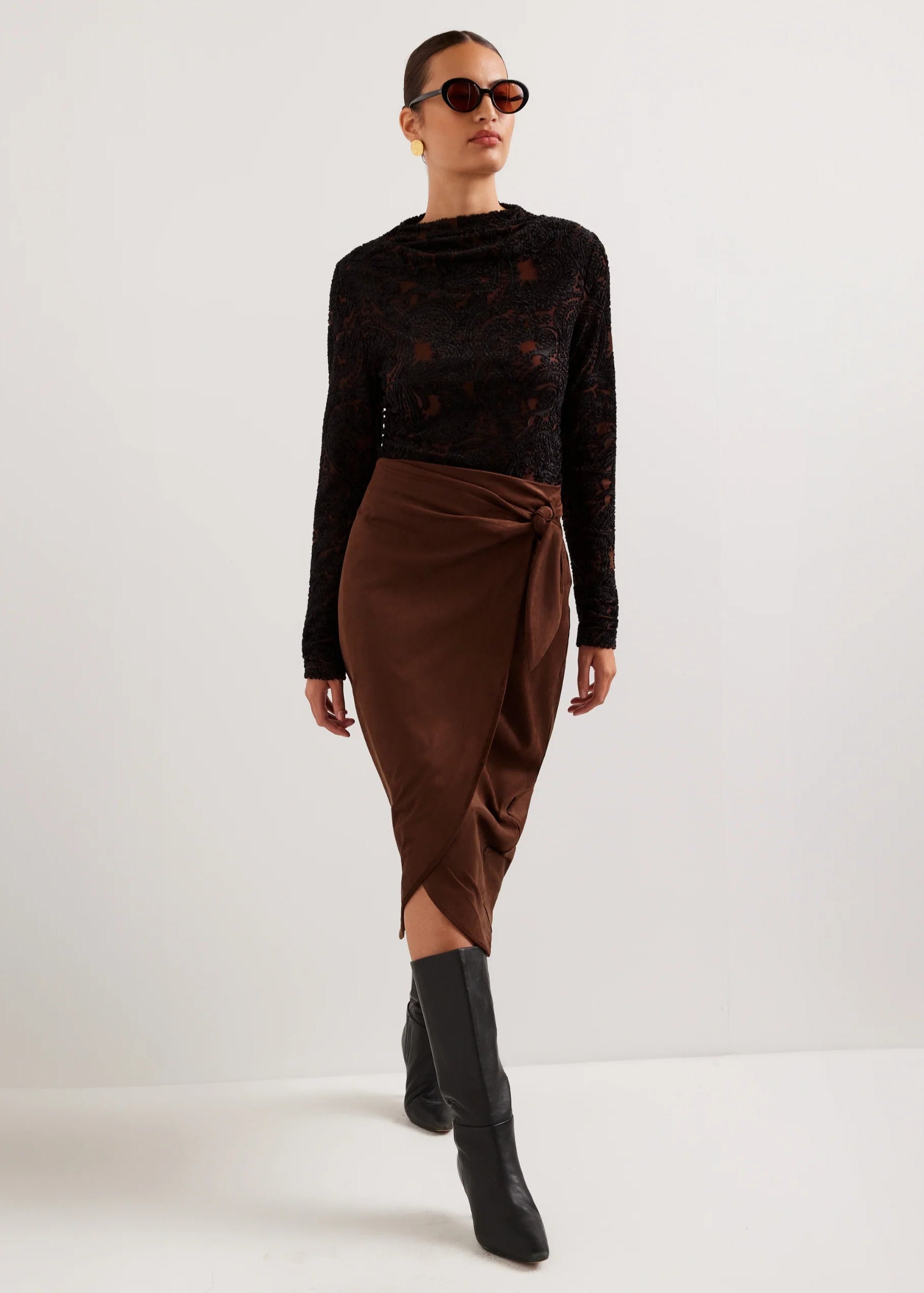A model wearing a black long-sleeve lace top and a brown wrap-style midi skirt, paired with black knee-high boots and sunglasses, standing against a plain light gray studio background.