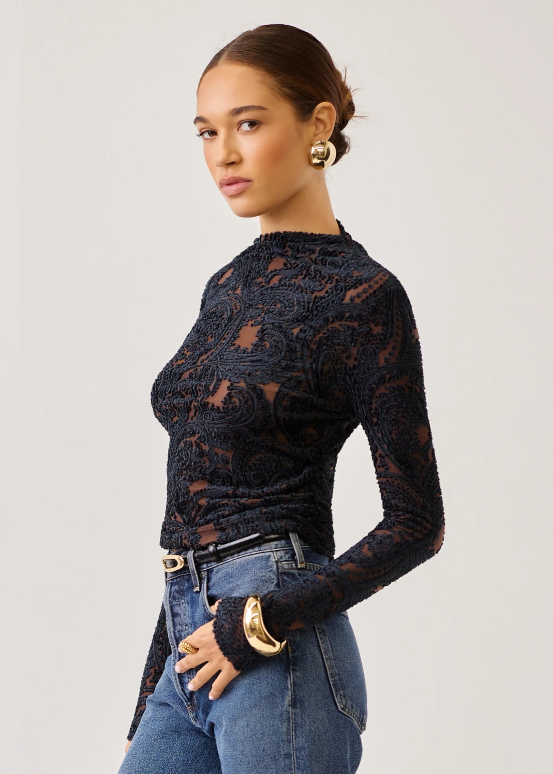 A woman modeling a black long-sleeve lace mock-neck top paired with blue jeans and gold jewelry, photographed against a plain light gray studio background.