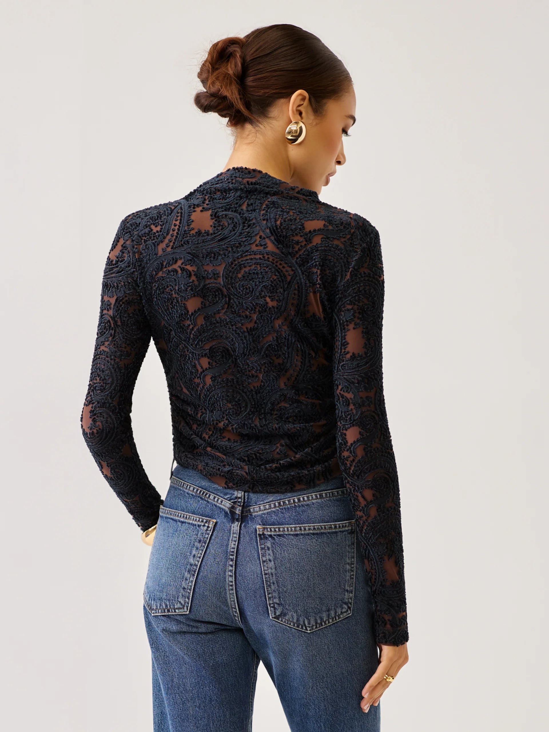 A woman shown from the back wearing a black lace long-sleeve top and blue jeans against a light gray studio background.