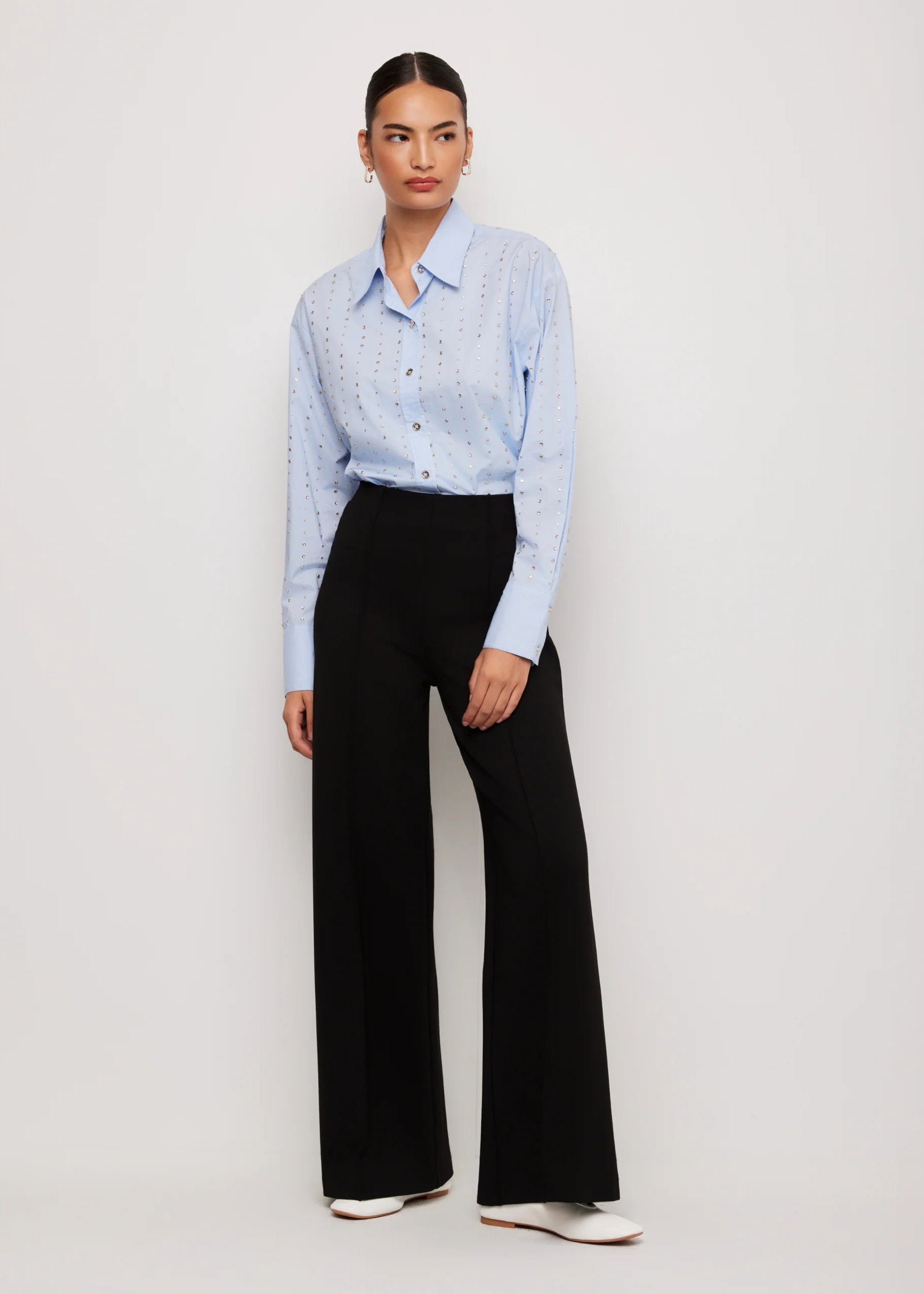 A model stands against a light gray studio background wearing a light blue long-sleeve button-up shirt with a subtle dotted pattern and black wide-leg trousers.