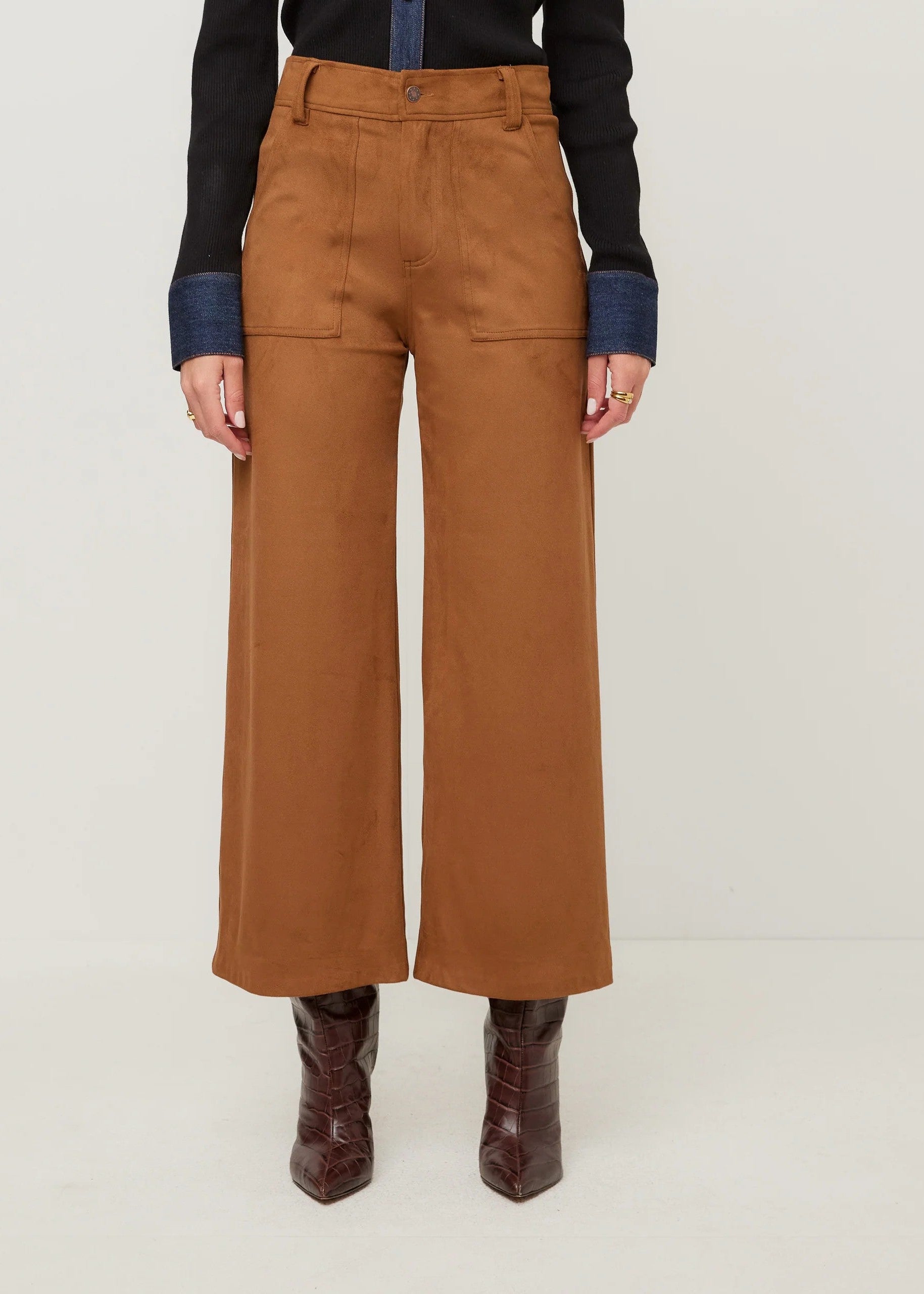 A model is shown from the waist down wearing a pair of wide-leg cropped trousers in a warm camel brown color, styled with a black and navy shirt and dark brown boots against a plain light background.