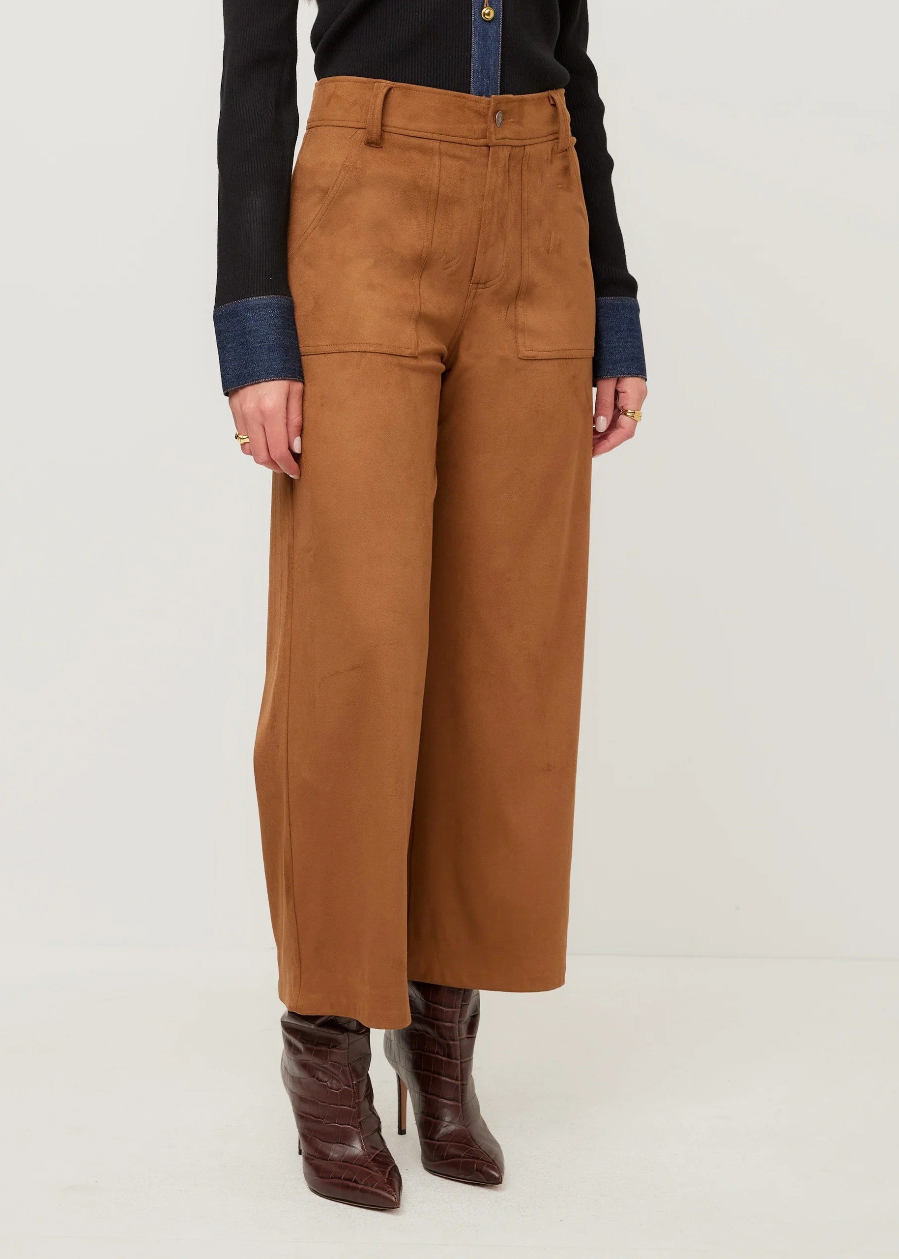A model wearing high-waisted, wide-leg brown trousers with a textured suede-like finish, styled with a black and navy top and dark brown ankle boots against a light gray studio background.