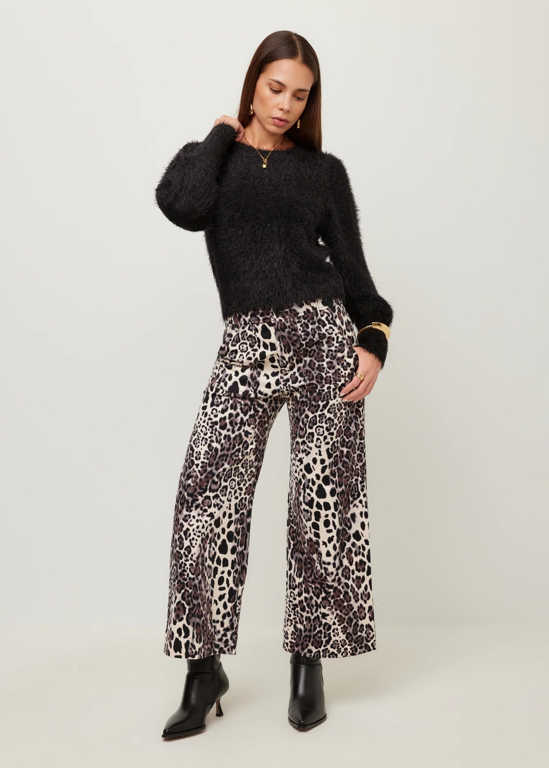 A woman modeling a black fuzzy long-sleeve sweater and wide-leg leopard-print pants with black pointed-toe ankle boots, photographed against a plain light gray studio background.