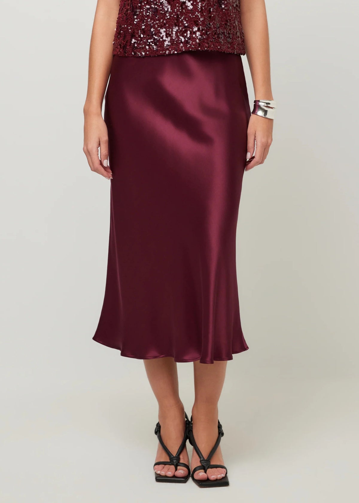 A woman models a satin midi skirt in deep burgundy, paired with a sequined top and black strappy heels, against a light gray studio background.