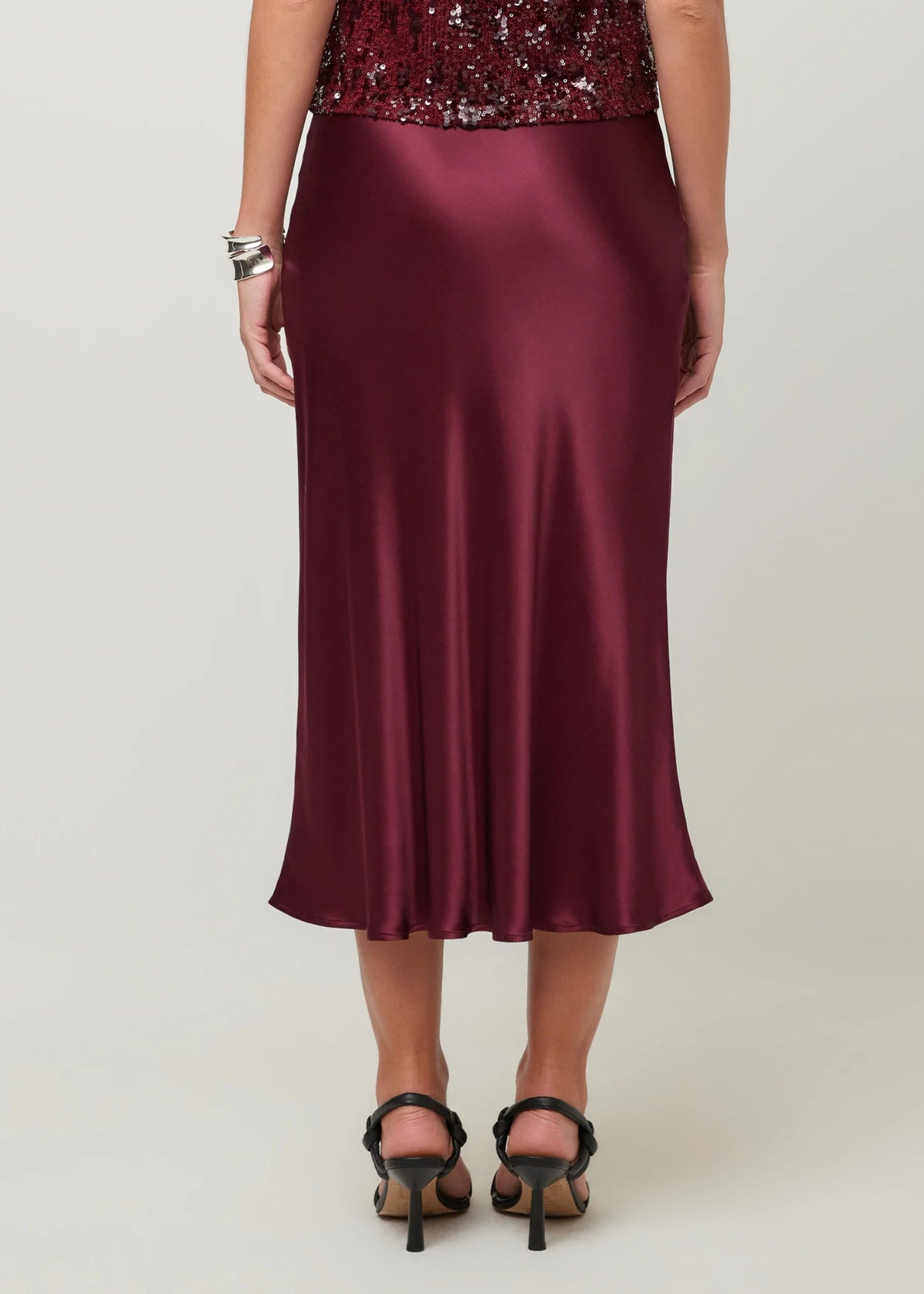 A woman shown from the back wearing a satin midi skirt in deep burgundy, paired with black high-heeled sandals and a silver bracelet, against a light gray studio background.