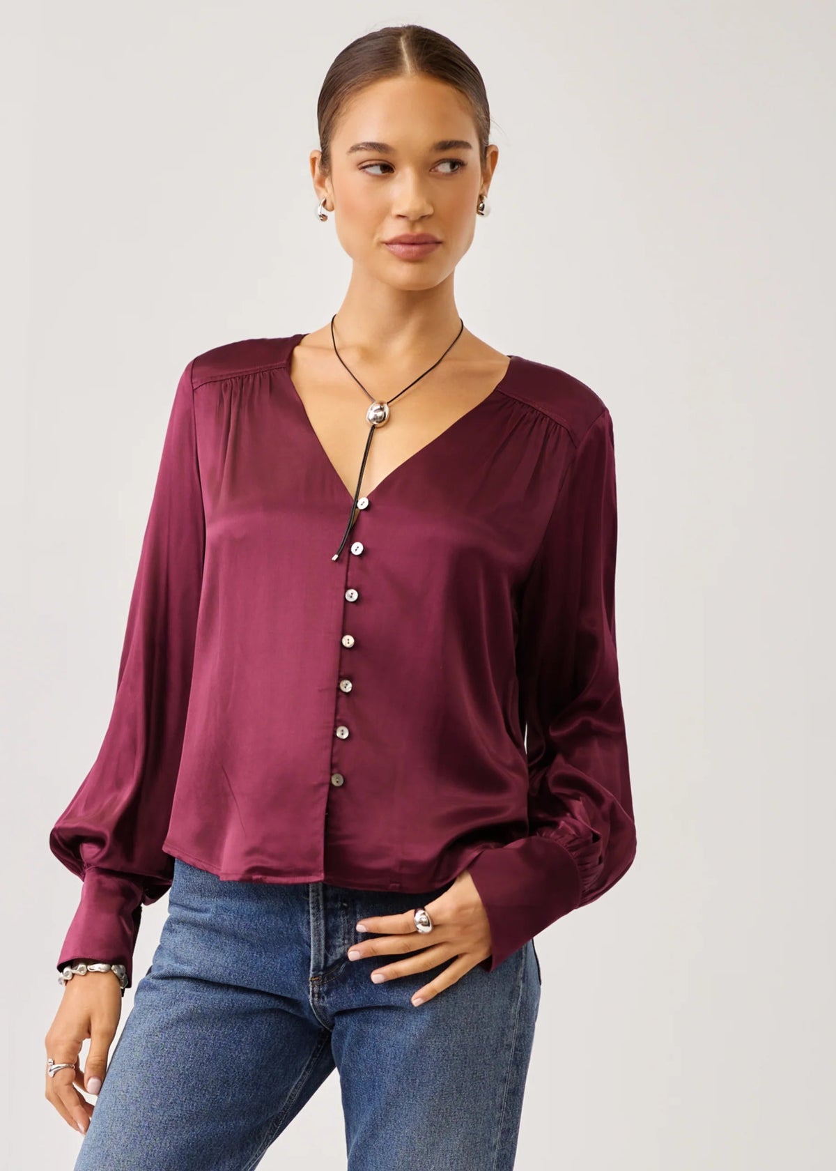 A woman modeling a long-sleeve satin blouse in deep burgundy with a button-down front and gathered neckline, paired with blue jeans against a light gray studio background.