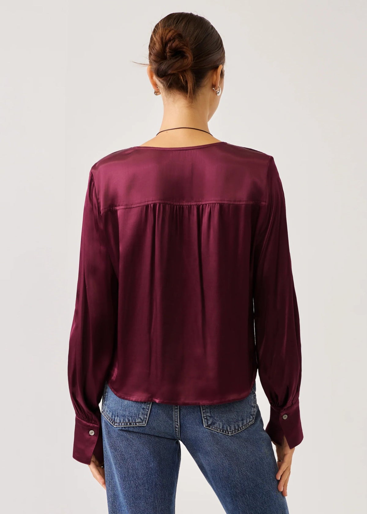 A woman viewed from the back wearing a long-sleeve satin blouse in deep burgundy and blue jeans, set against a plain light gray studio background.