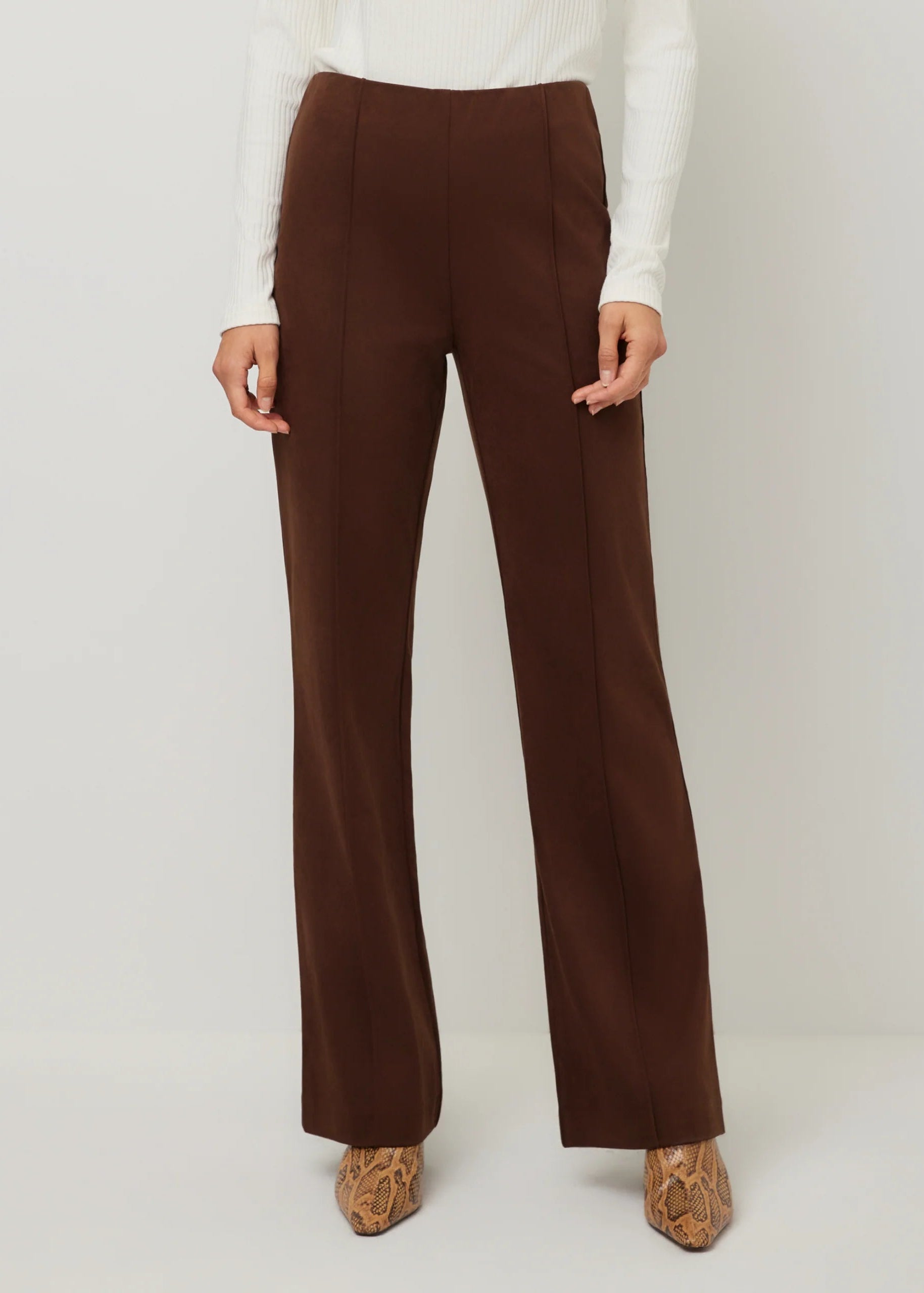 A model wearing high-waisted, wide-leg brown trousers, paired with a white ribbed long-sleeve top and brown snakeskin-patterned shoes, against a light gray studio background.