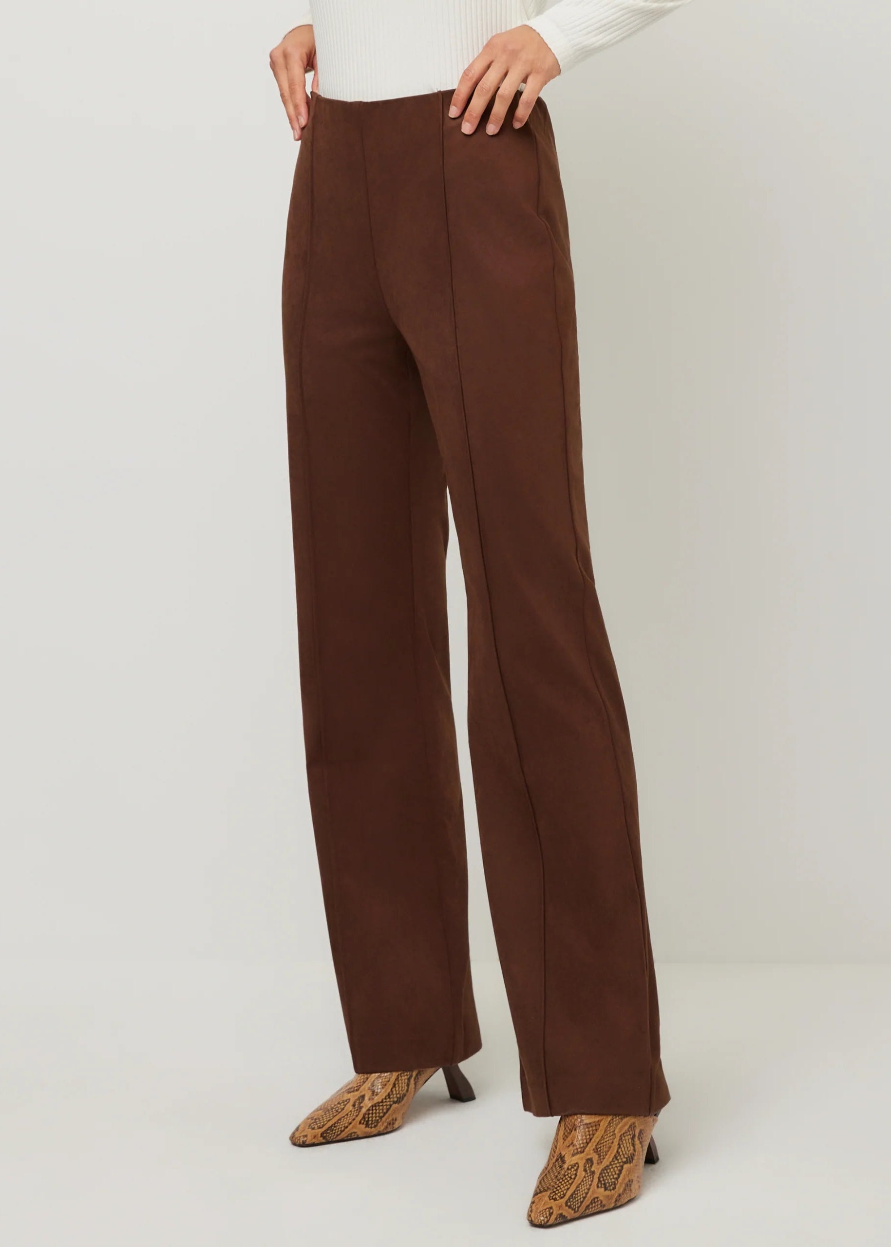 A model wearing high-waisted, wide-leg brown trousers paired with a white top and brown snakeskin-patterned heels, shown from the waist down against a light gray studio background.