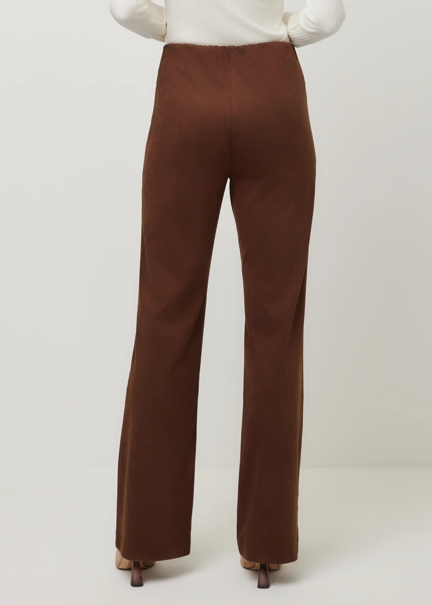 A person shown from the back wearing brown wide-leg trousers and a white ribbed top, standing against a plain light gray studio background.