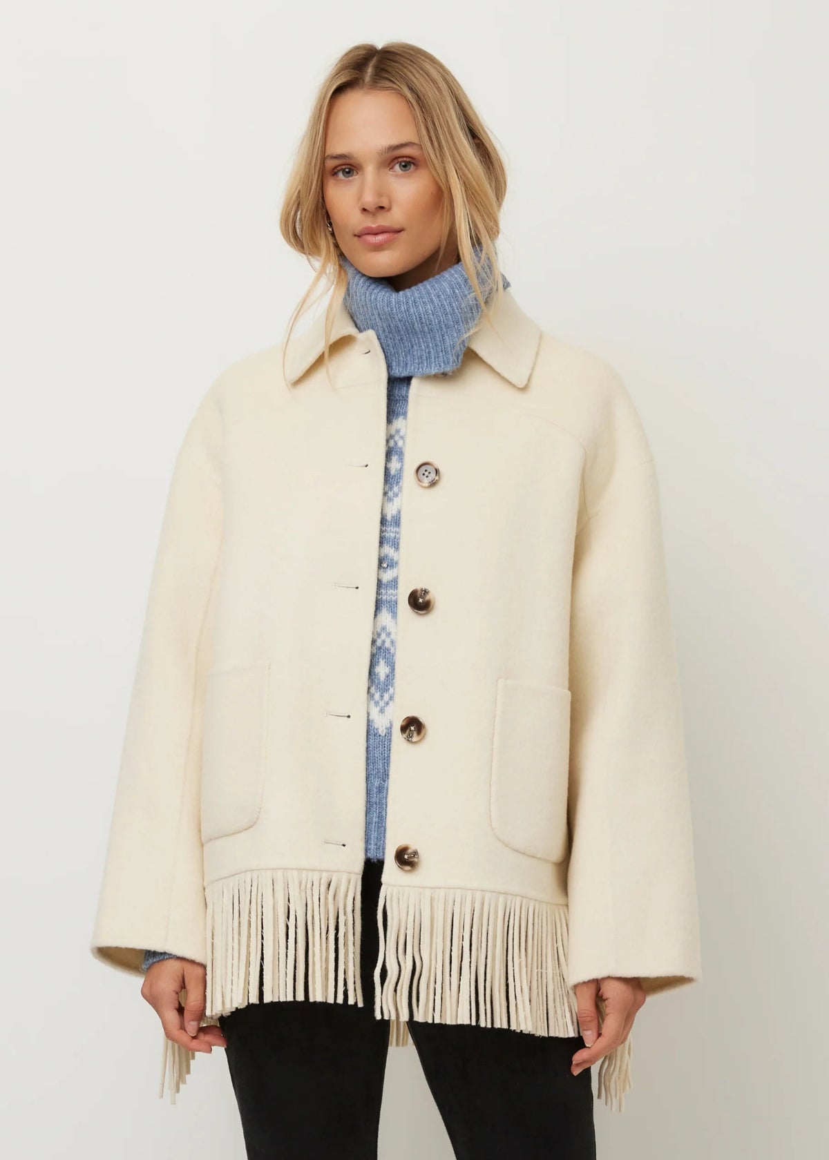 A woman models a cream-colored wool coat with fringe detailing and large front pockets, layered over a blue knit turtleneck sweater and black pants, against a plain light gray background.