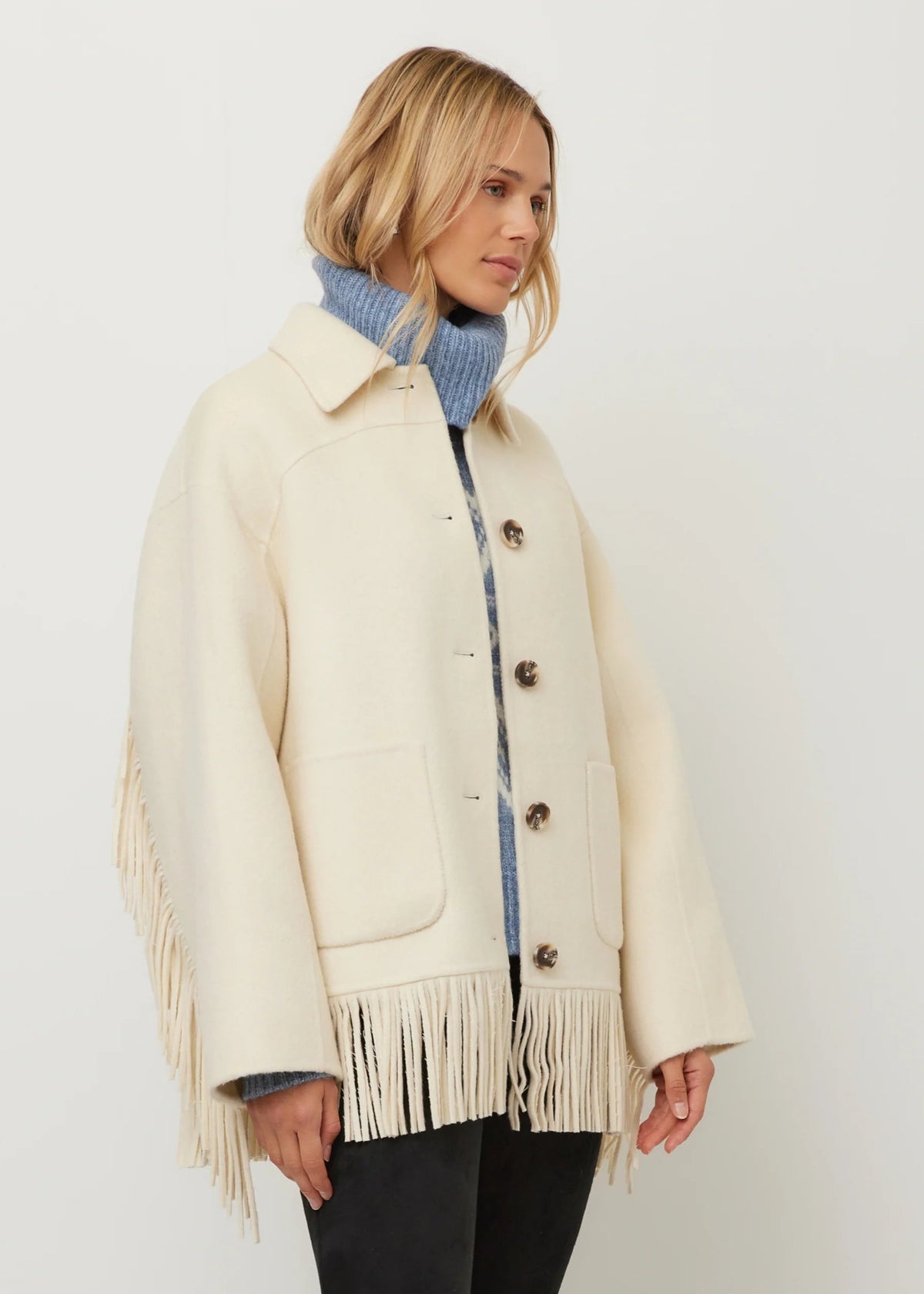 A woman wearing a cream-colored fringed jacket over a blue knit turtleneck sweater and black pants, photographed against a plain light gray studio background.