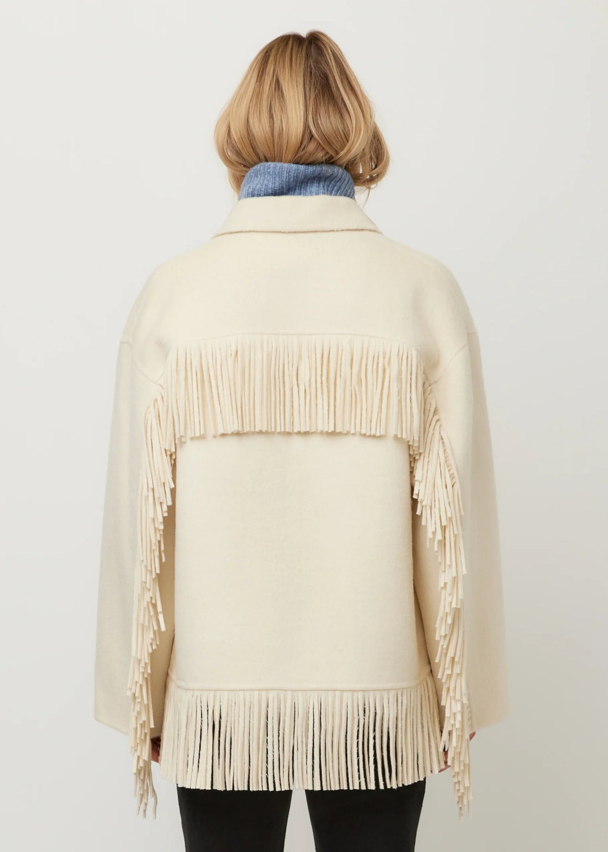 A woman viewed from the back wearing a cream-colored fringed jacket over a blue turtleneck sweater and black pants, set against a plain light gray studio background.