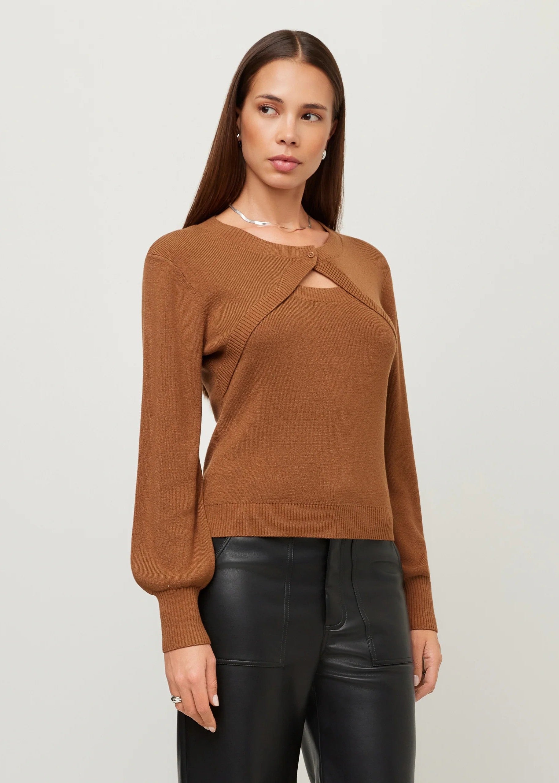 A woman modeling a brown knit sweater with a unique asymmetrical crossover front and a small chest cutout, paired with black leather-look pants against a light gray studio background.