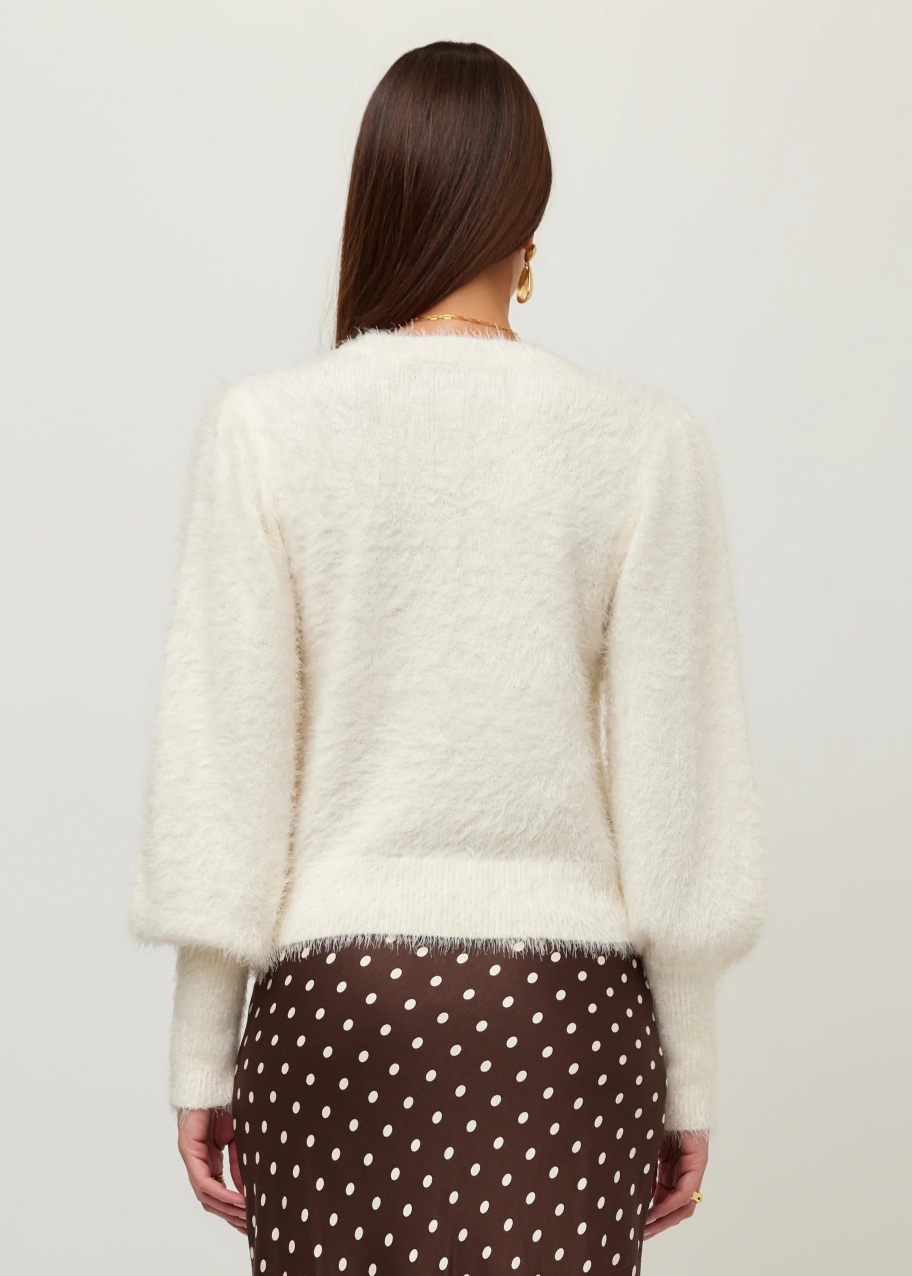 A woman shown from the back wearing a fuzzy off-white cropped sweater and a brown skirt with white polka dots, against a plain light gray studio background.