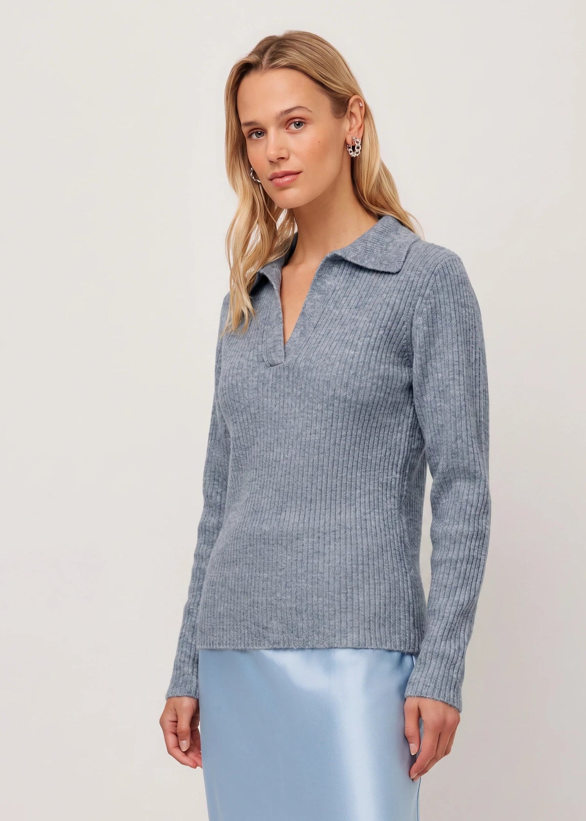 A woman modeling a light blue-gray ribbed knit polo sweater with a collar and split neckline, paired with a light blue satin skirt, against a plain off-white studio background.