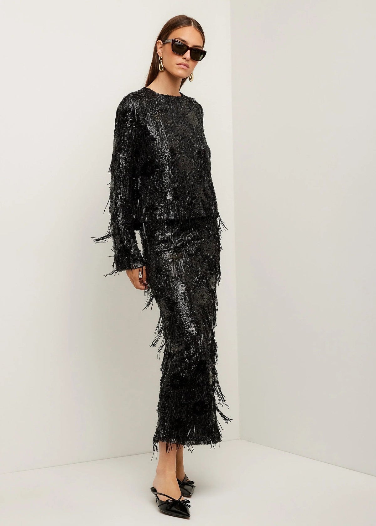 A model wearing a black long-sleeve top and matching fringed maxi skirt set, styled with black sunglasses and black pointed flats, standing in a minimalist white studio corner.