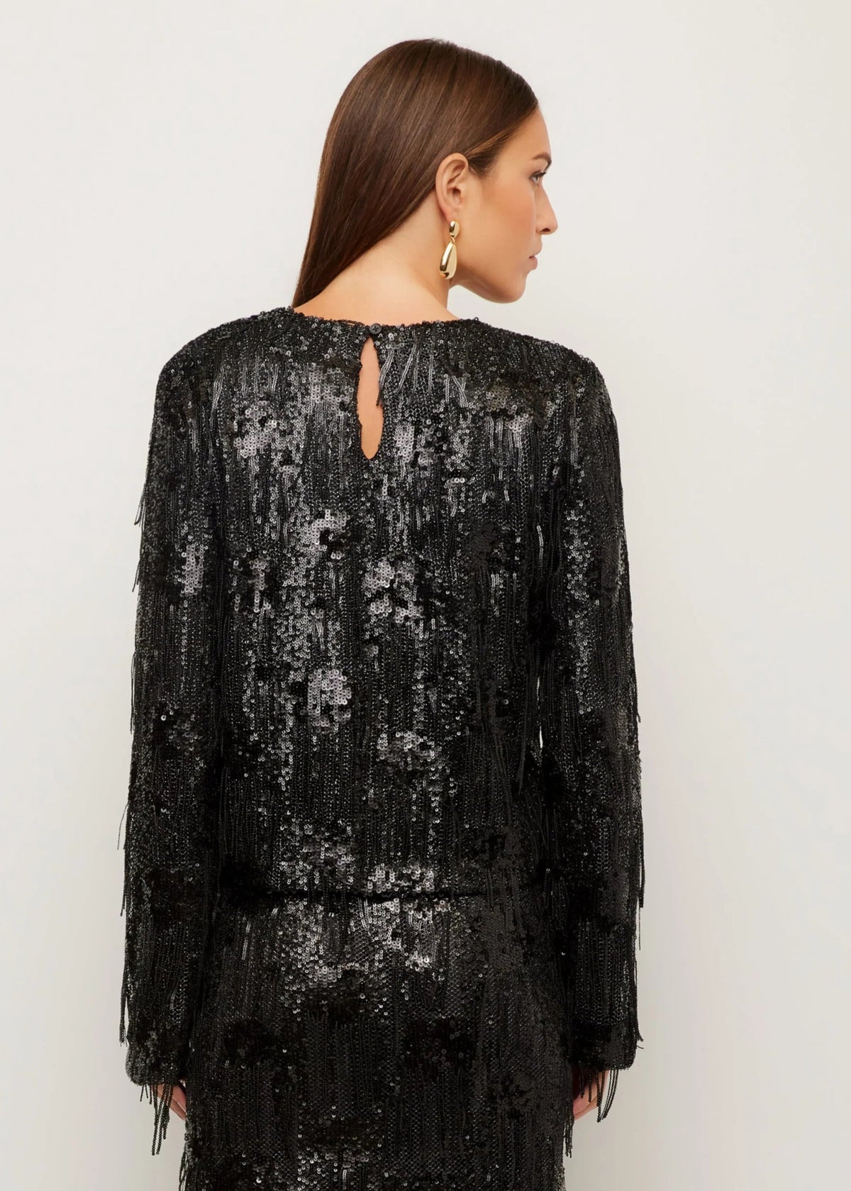 A woman shown from the back wearing a black long-sleeve fringed dress with sequined vertical detailing and a small keyhole cutout at the neckline, against a plain light gray studio background.