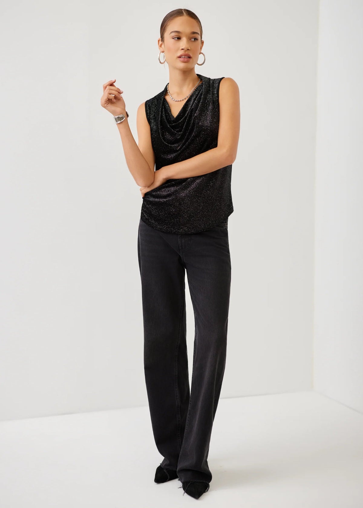 A model stands in a studio wearing a black sleeveless draped top with a subtle glitter texture and black wide-leg trousers.