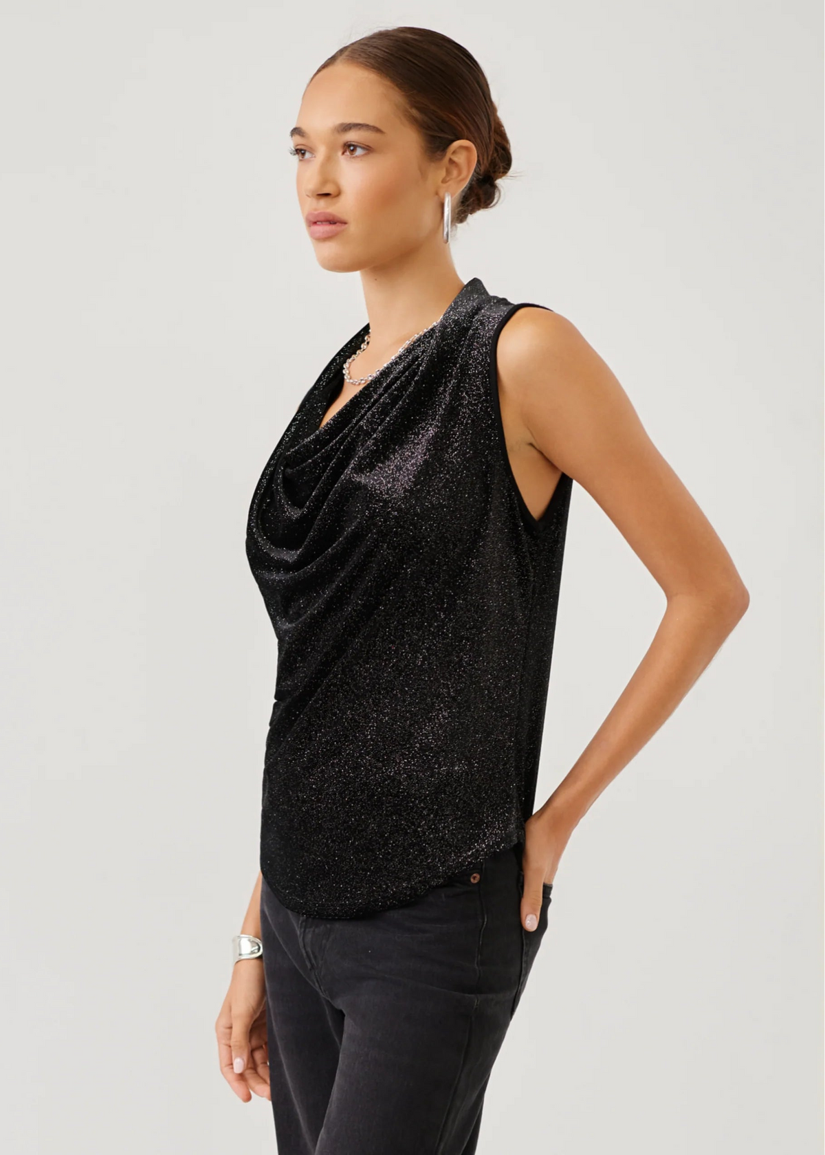 A woman modeling a sleeveless black draped cowl-neck top with a subtle glittery texture, paired with dark pants, against a light gray studio background.