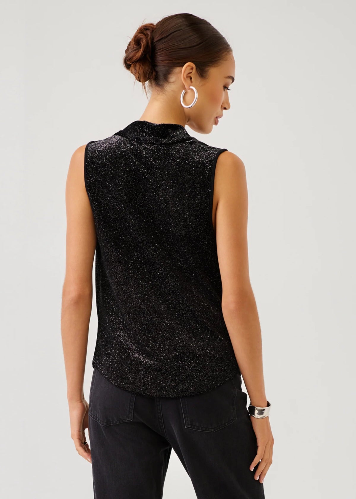A woman shown from the back wearing a sleeveless black textured top with a mock neck and black pants, photographed against a light gray studio background.