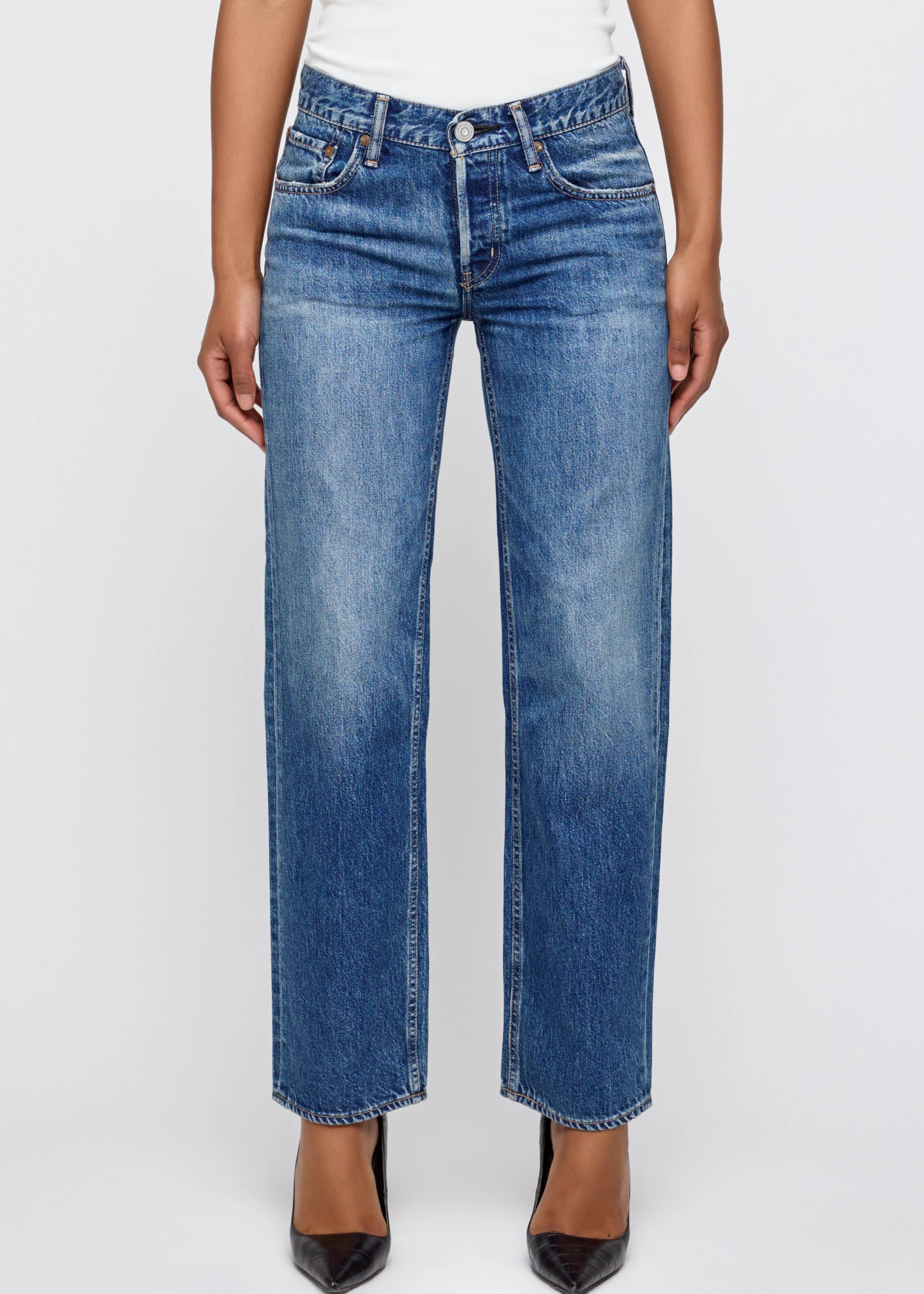 A person wearing medium blue straight-leg denim jeans with whiskering and subtle fading, paired with a white top and black pointed-toe heels, against a light gray studio background.