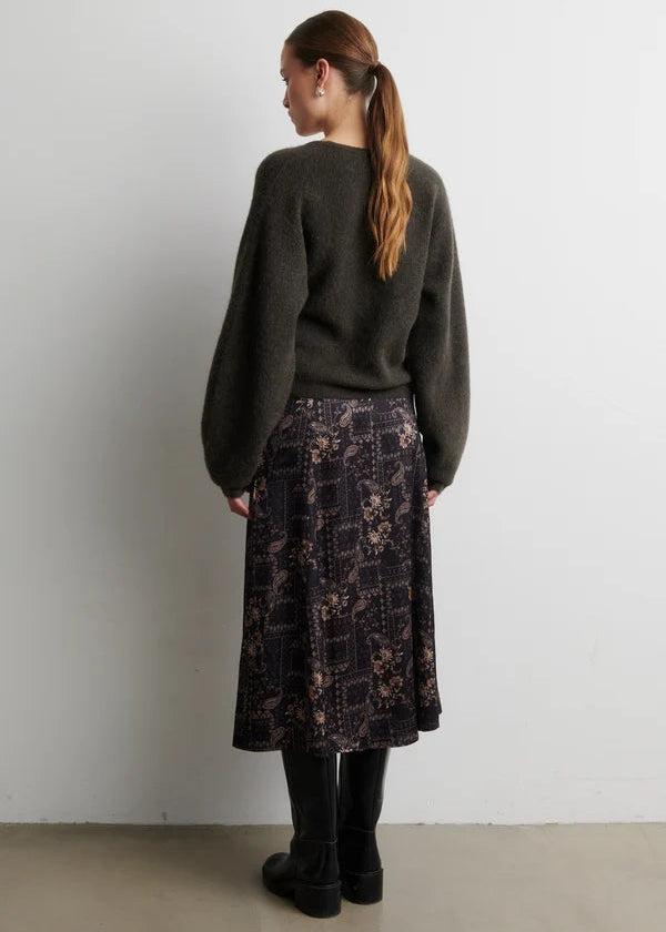 A woman stands with her back to the camera wearing a dark olive green knit sweater, a dark patterned midi skirt, and black boots, set against a plain light gray wall.