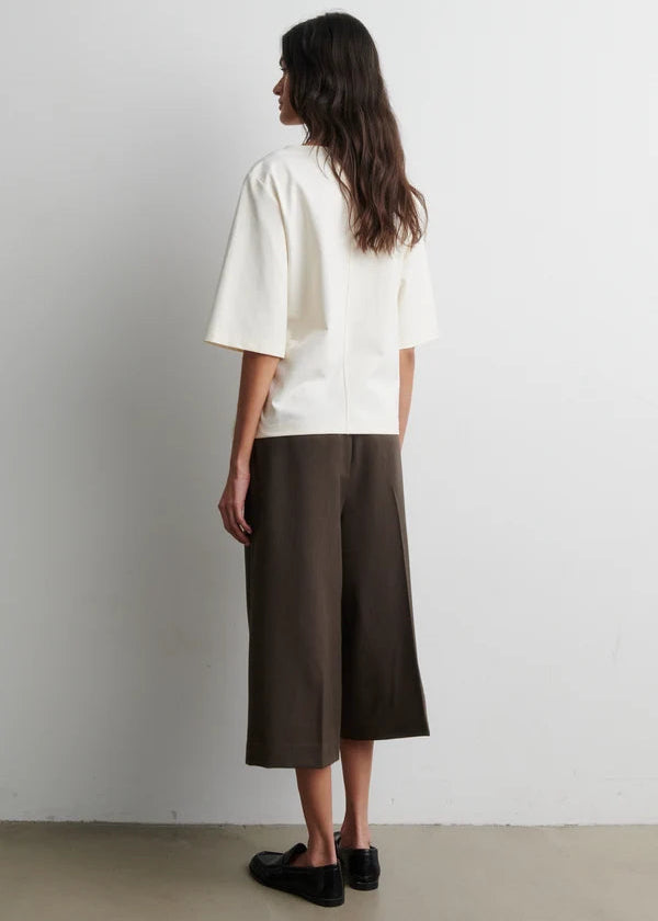 Woman wearing a white shirt and brown pants standing against a plain white wall.