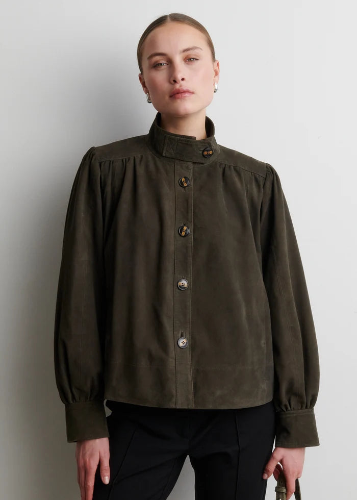 A woman wearing a dark olive brown long-sleeve button-up shirt with a high gathered collar, paired with black trousers, standing against a light gray wall.
