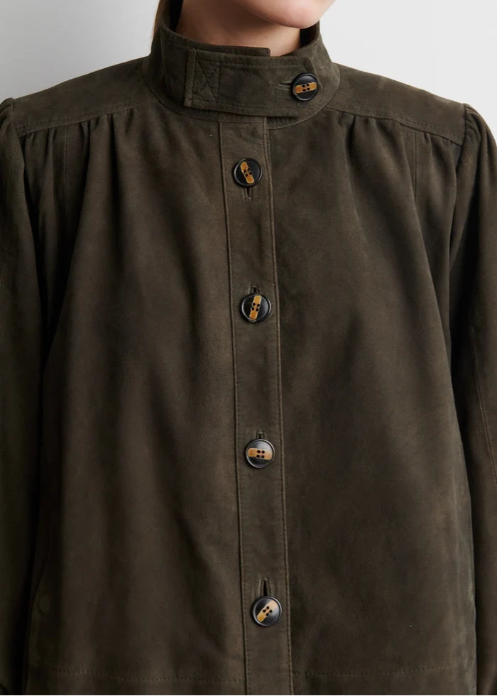 Close-up of a person wearing a dark olive brown suede jacket with a high stand-up collar and decorative brown buttons with gold detailing.