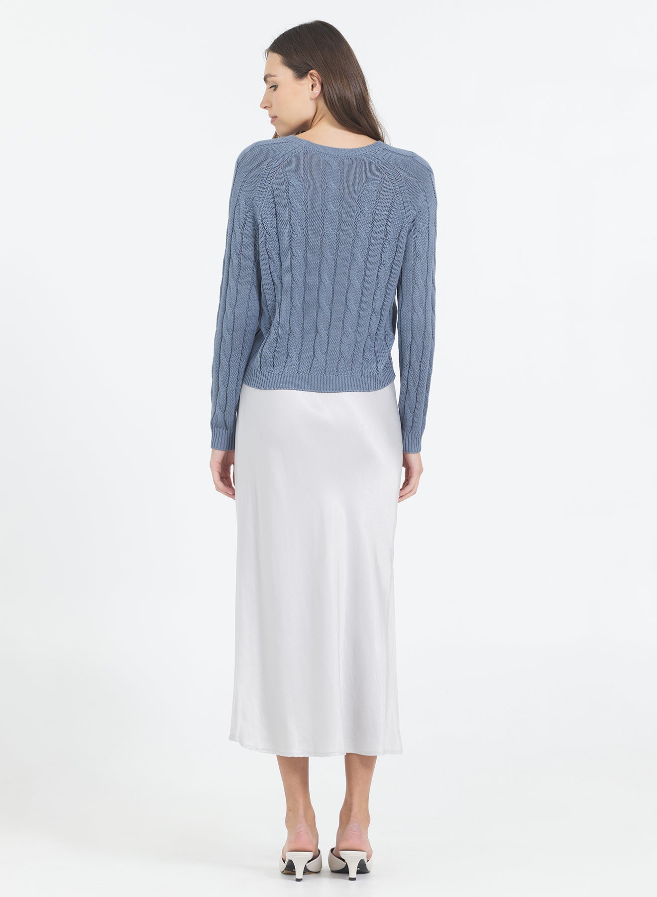 Woman wearing the One Grey Day blue Raleigh Cable sweater in Cadet and white skirt on a white background wearing a blue sweater 
