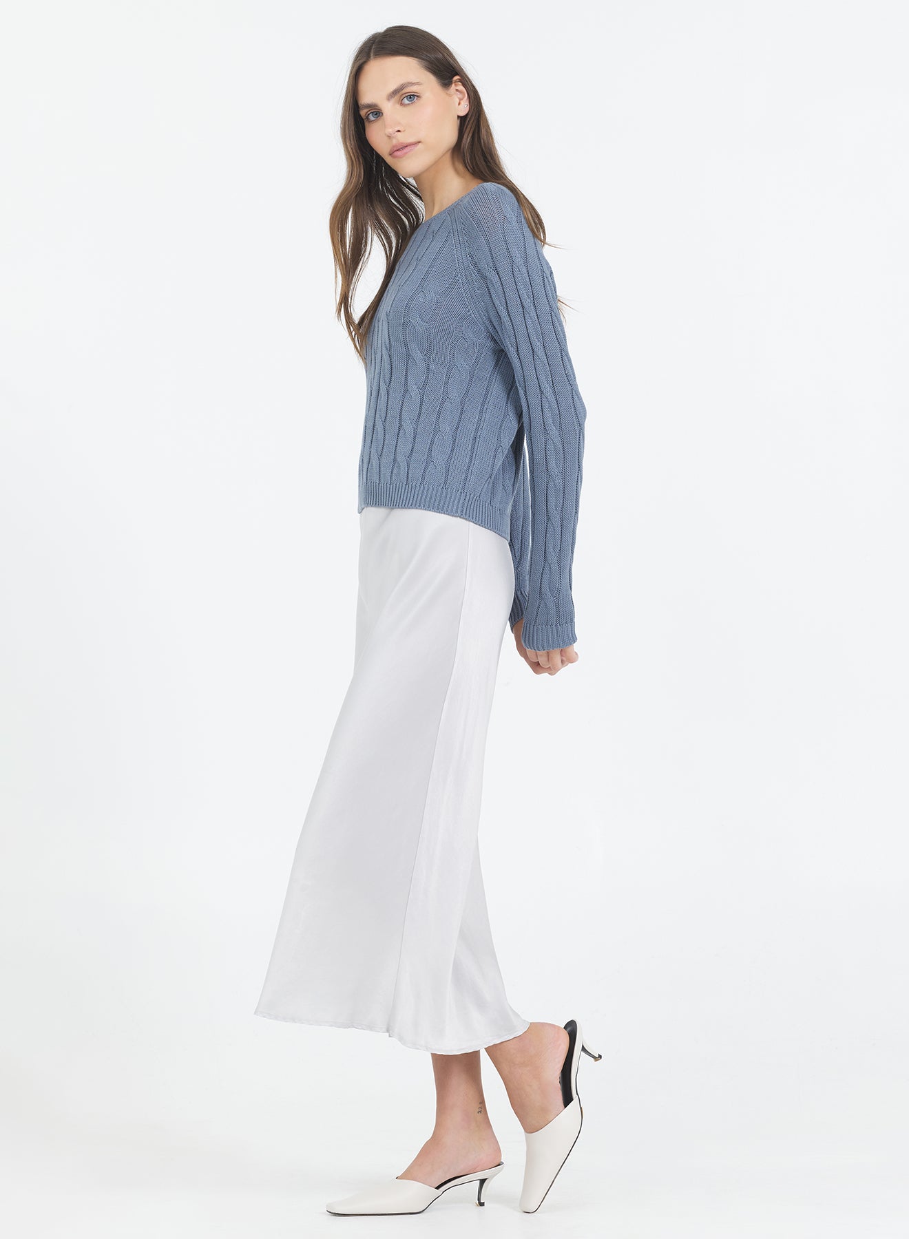 Woman wearing the One Grey Day blue Raleigh Cable sweater in Cadet and white skirt on a white background