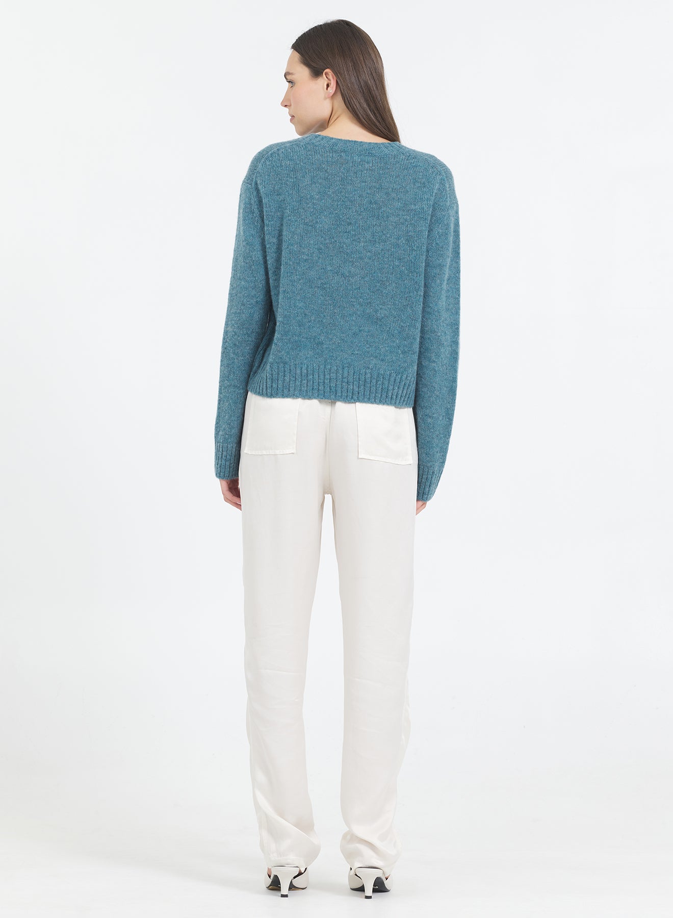 Woman wearing a One Grey Day teal lagoon Levanzo sweater and white pants on a white background
