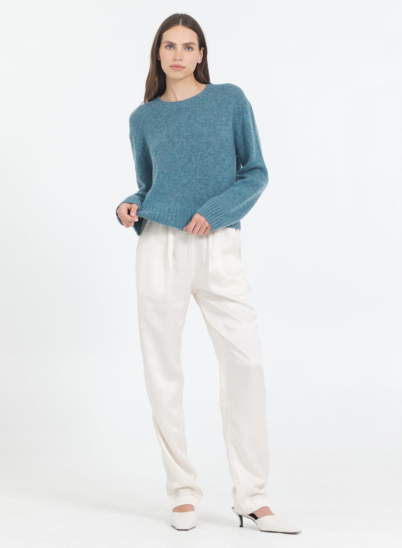 Woman wearing a blue lagoon One Grey Day Levanso sweater and white pants on a white background