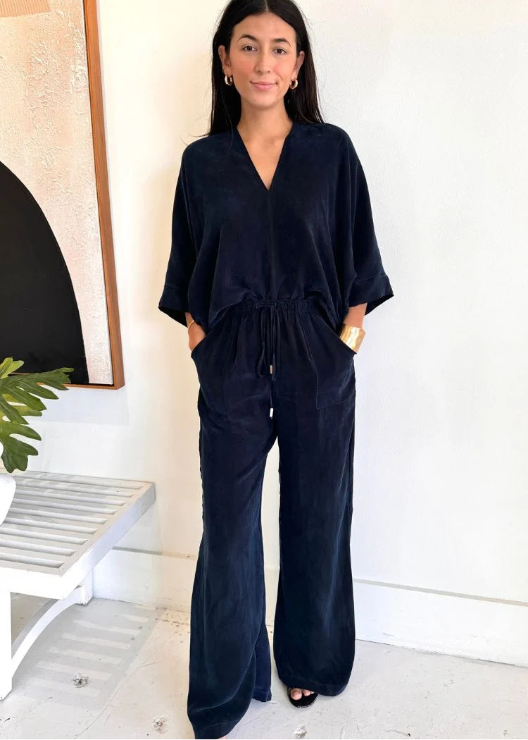 A woman modeling a dark navy blue velvet jumpsuit with wide legs, a V-neck, and a drawstring waist, standing in a brightly lit room with a white wall.