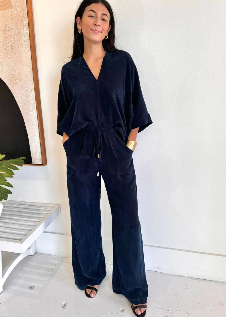 A woman modeling a dark navy blue velvet jumpsuit with wide legs, a V-neck, and a drawstring waist, standing in a brightly lit indoor setting.