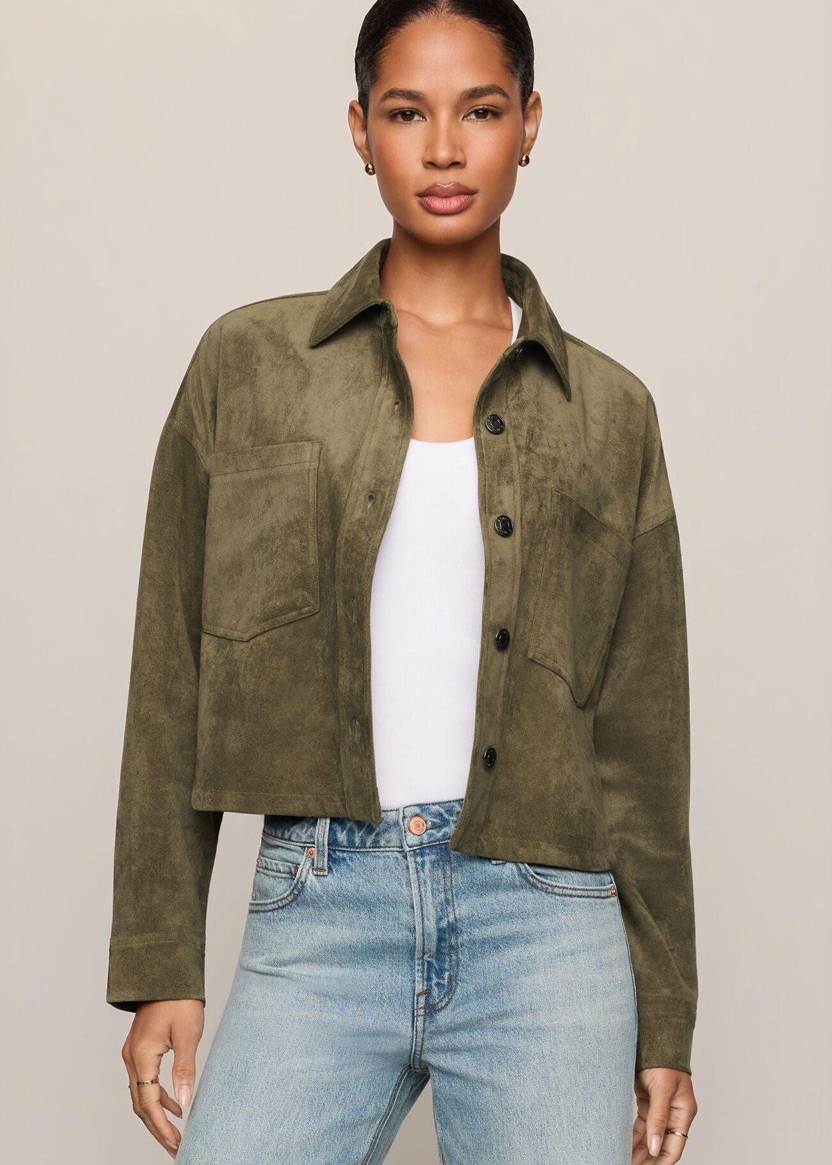 A woman modeling a cropped, long-sleeve olive green suede or nubuck button-up jacket over a white tank top and light-wash blue jeans, set against a solid light beige studio background.