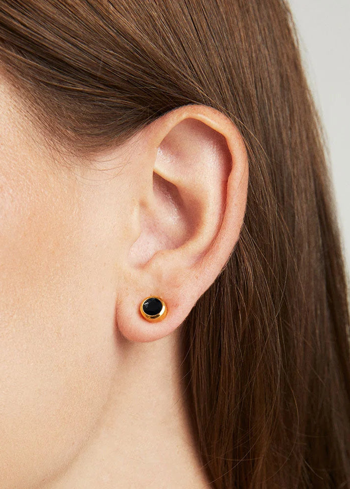 A close-up of a model's ear wearing a small gold hoop earring with a black center, against a light gray background.