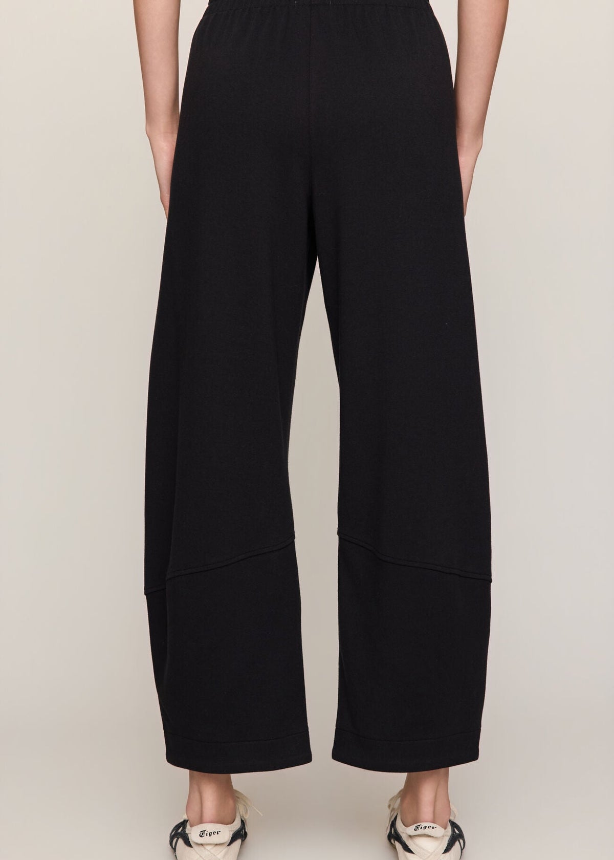A person shown from the back wearing loose black wide-leg cropped pants with an elastic waistband, paired with white sneakers, against a plain light gray studio background.