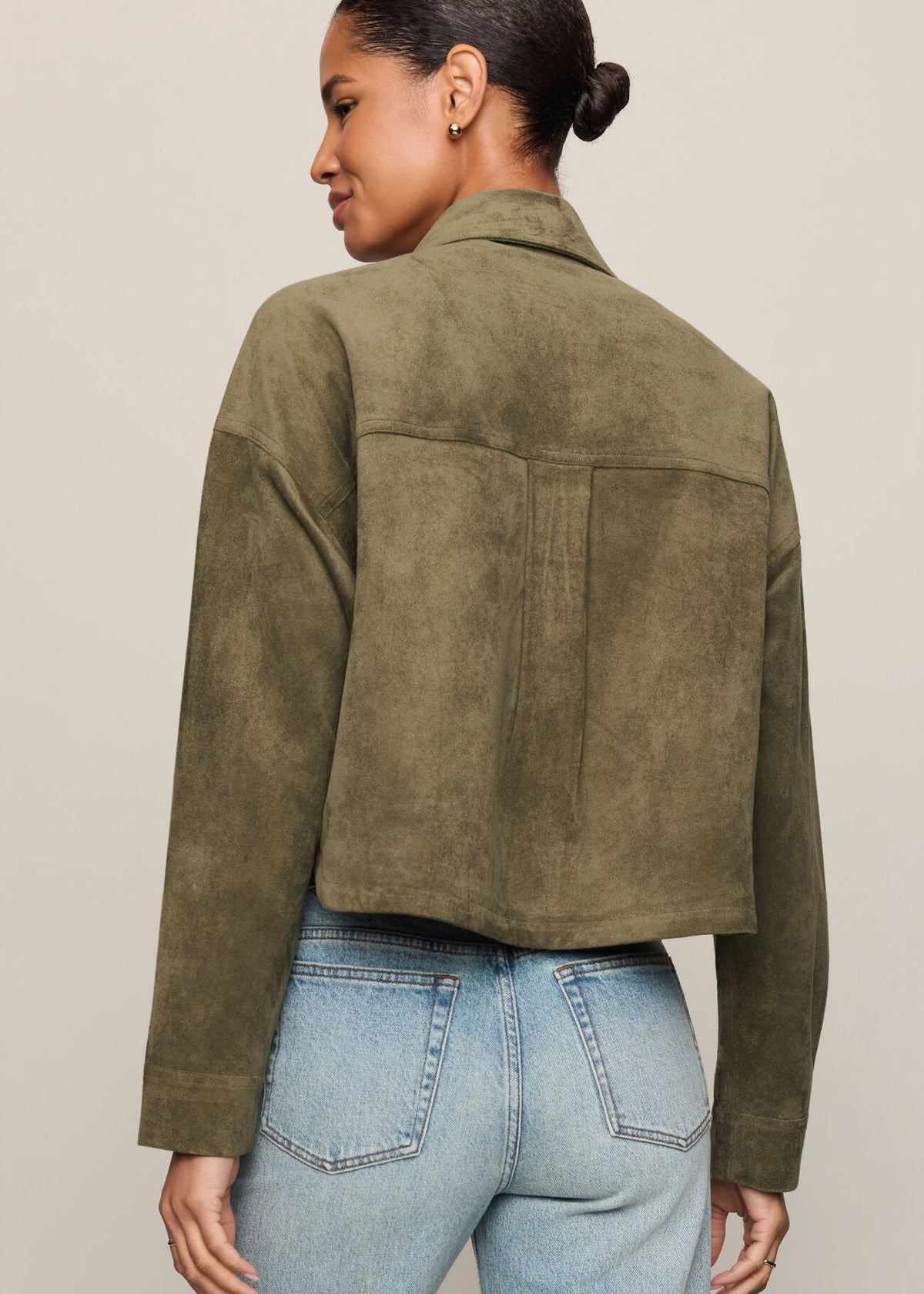 A woman shown from the back wearing an olive green cropped suede jacket and light blue jeans, photographed against a plain light beige studio background.