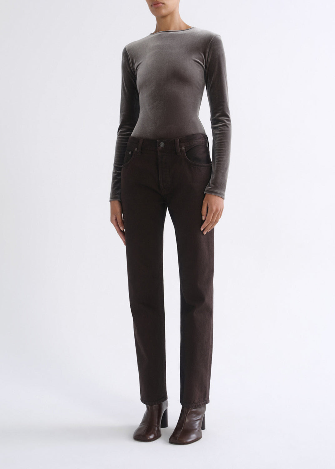 A model wearing a dark brown long-sleeve velvet top and dark brown straight-leg jeans, paired with dark brown heeled ankle boots, against a plain light gray studio background.